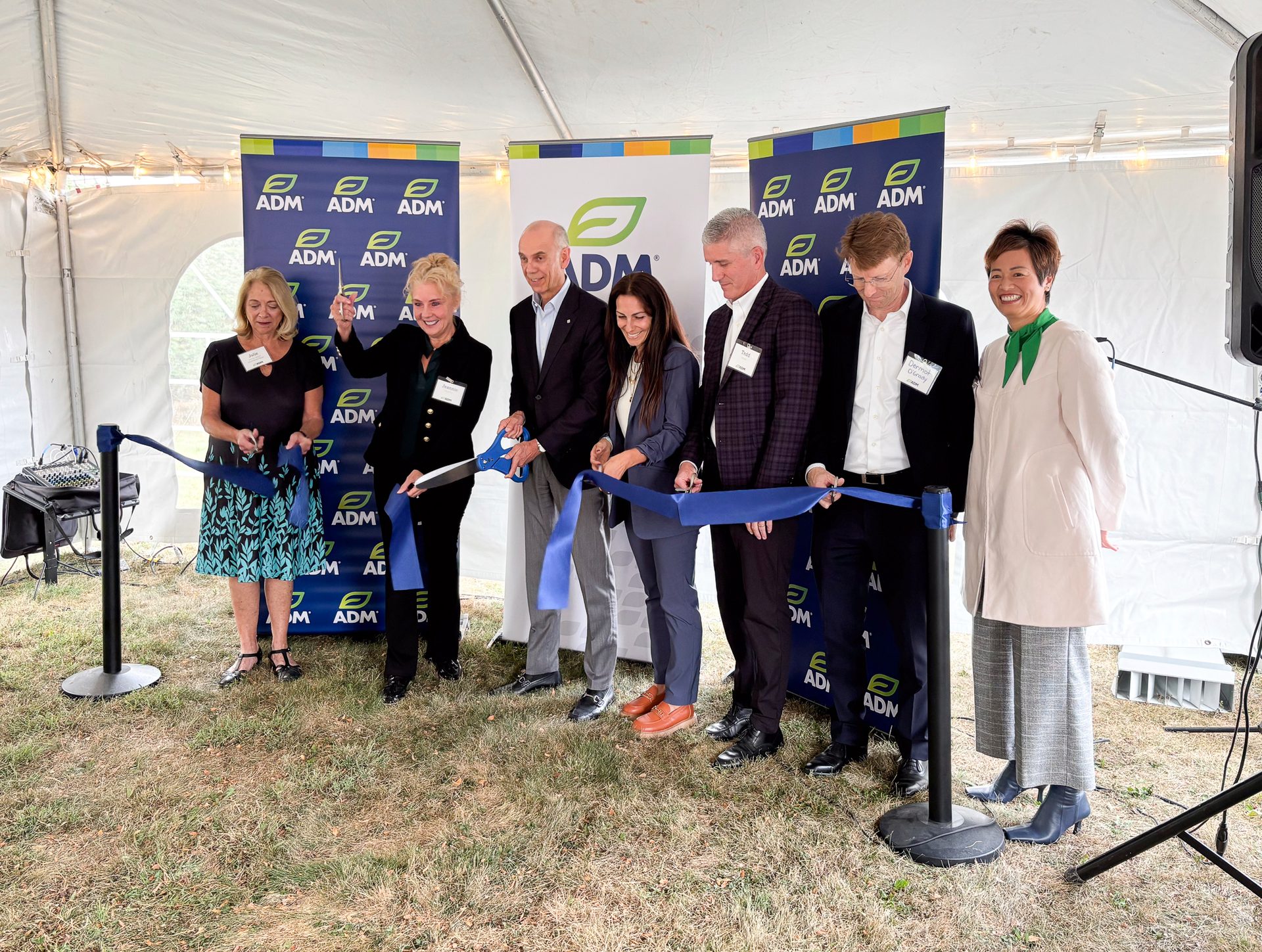 Seven people cut a blue ribbon at an outdoor ADM ceremony under a tent.
