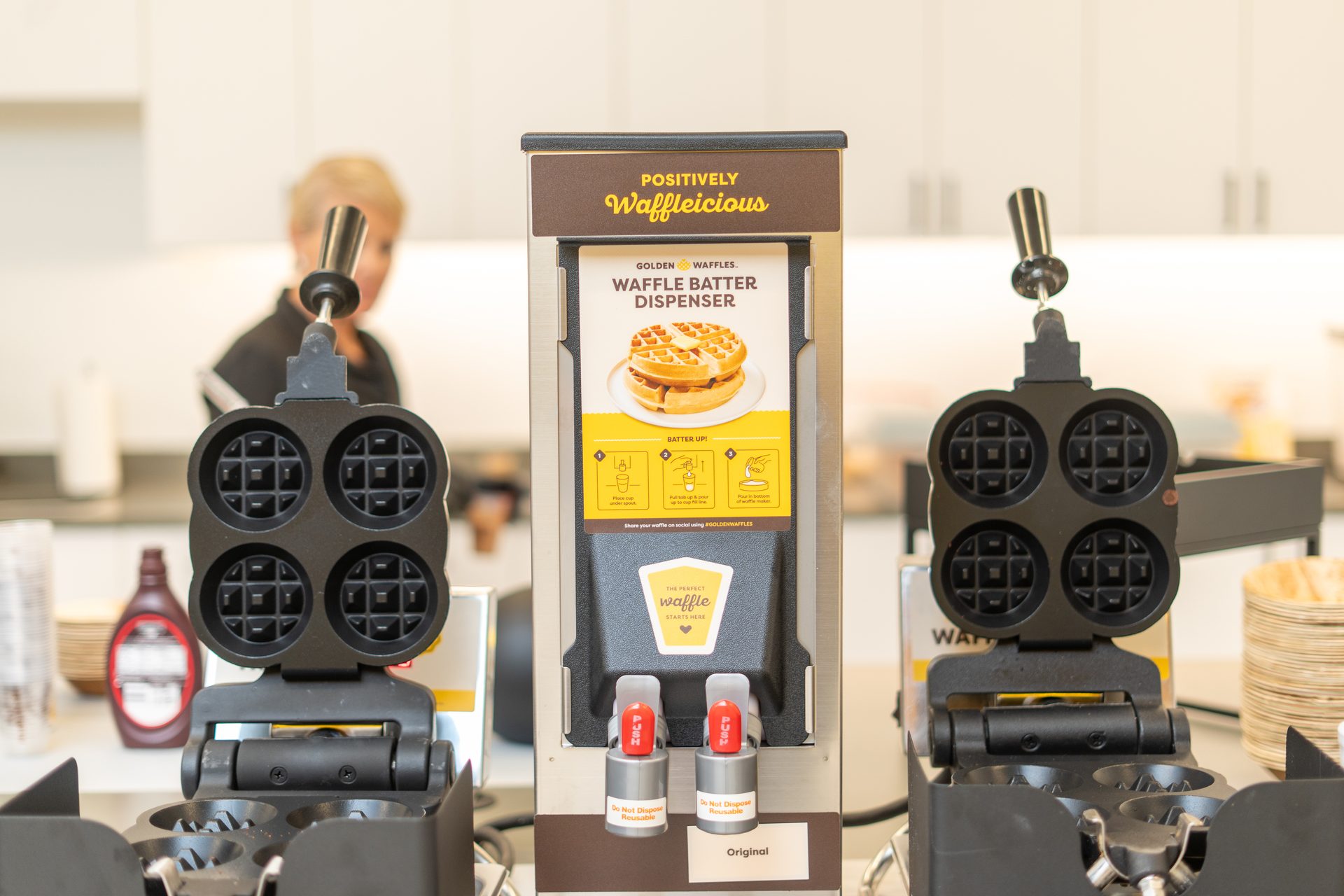 A 'Positively Waffleicious' waffle batter dispenser flanked by two waffle irons, with a person blurred in the background.