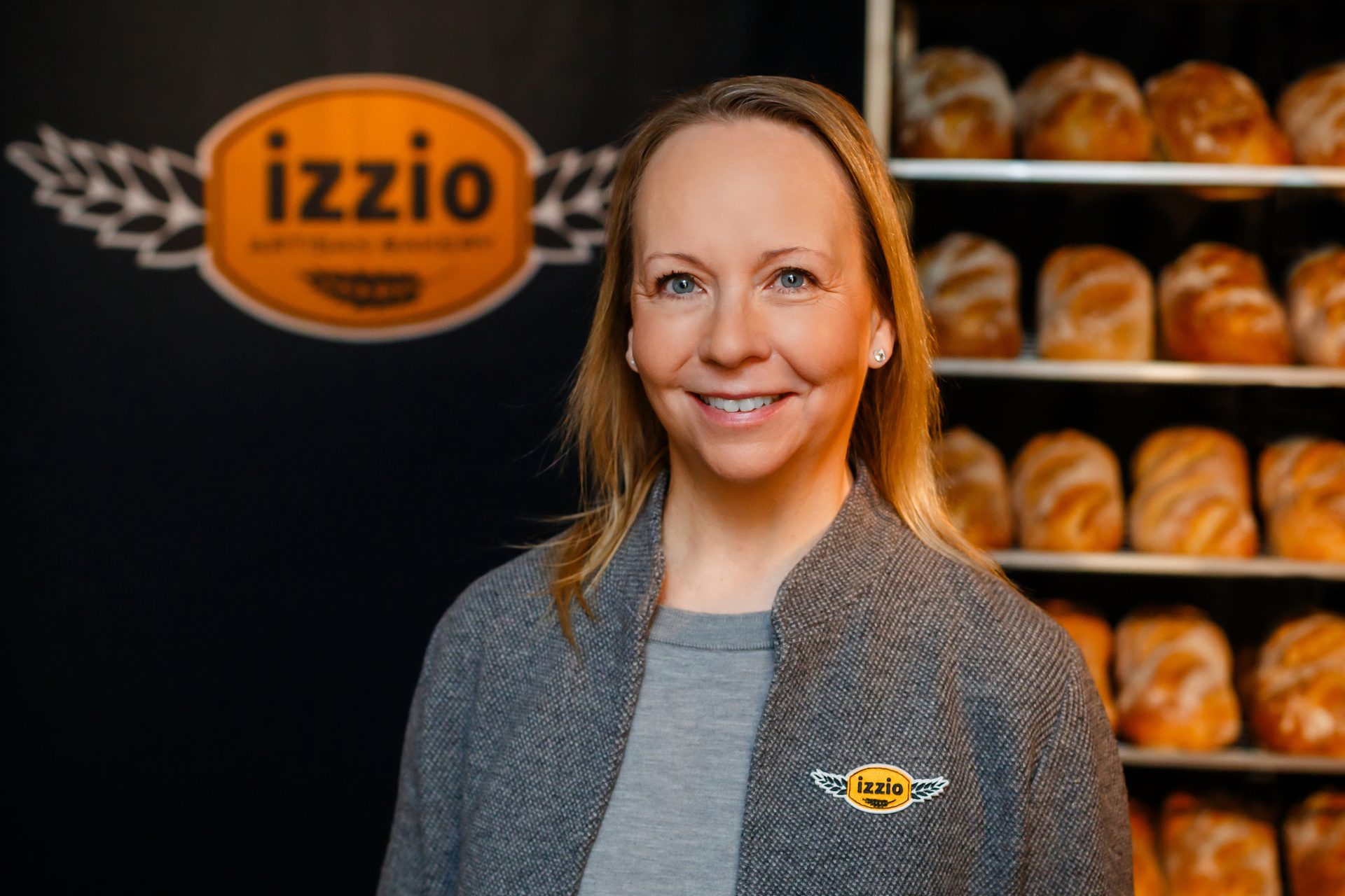 Smiling woman in gray jacket with Izzo logo, shelves of bread and brand sign in background.