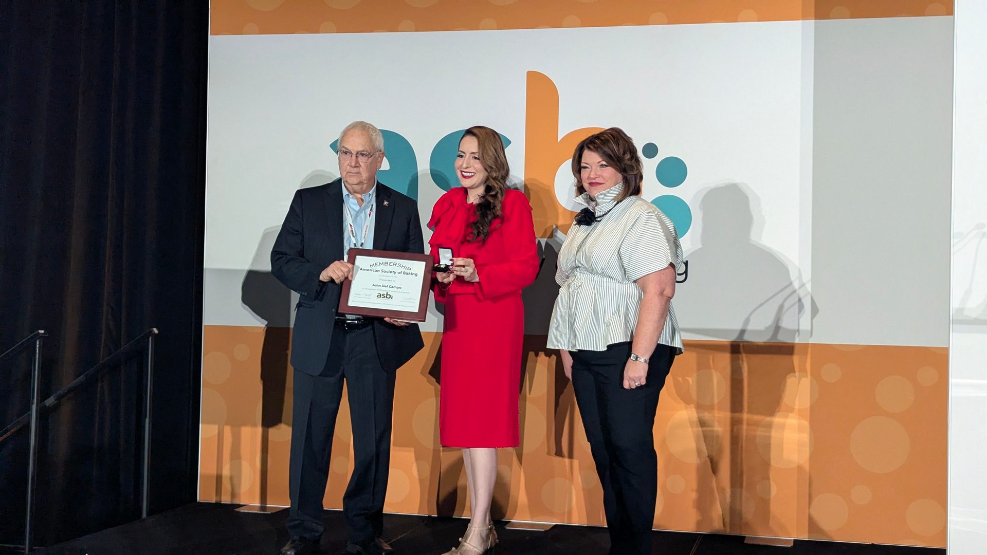 Man receives "American Society of Baking" award from woman in red, another woman stands by.