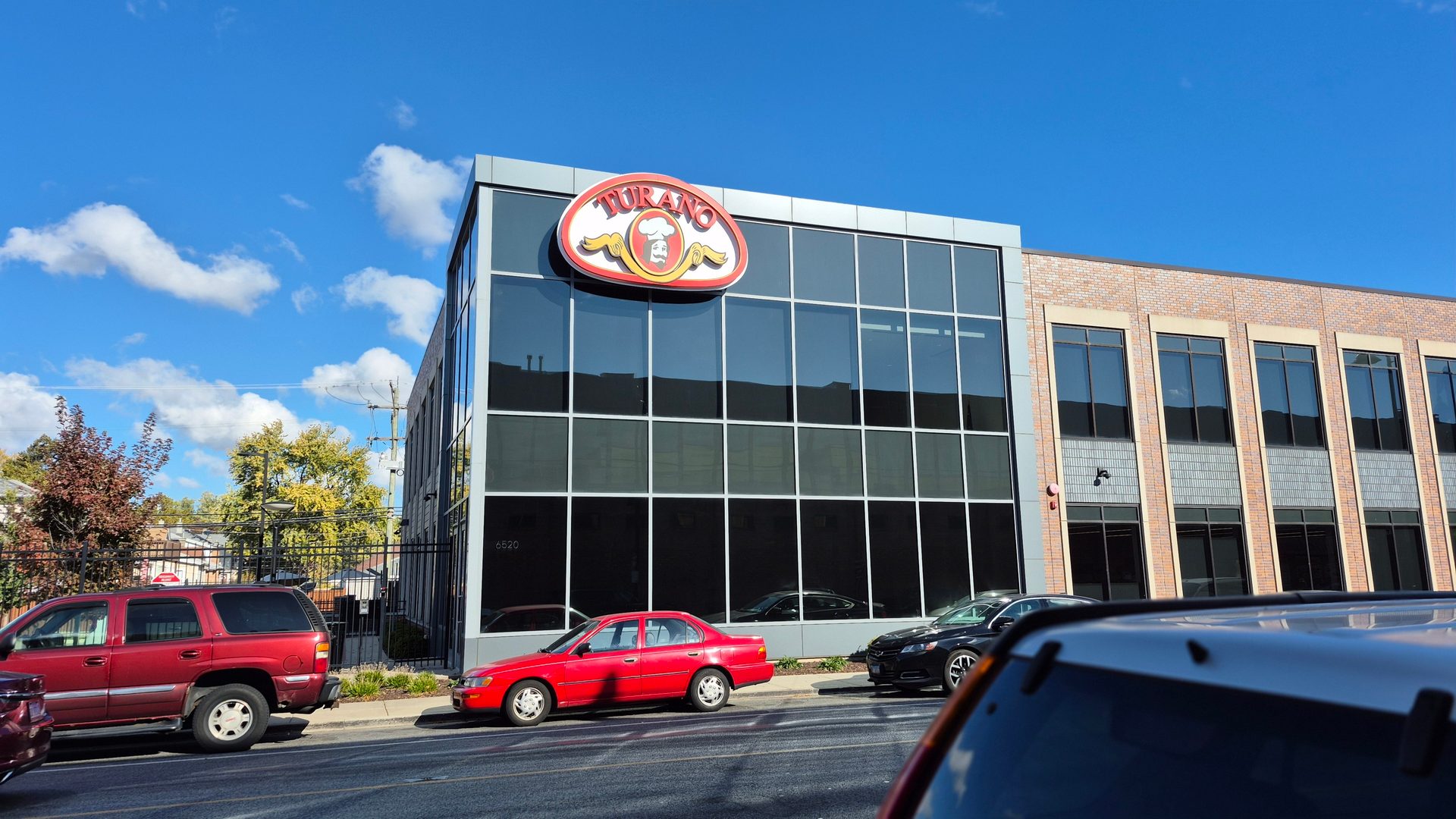 A modern building with a large "Turano" sign, cars parked outside, under a blue sky.