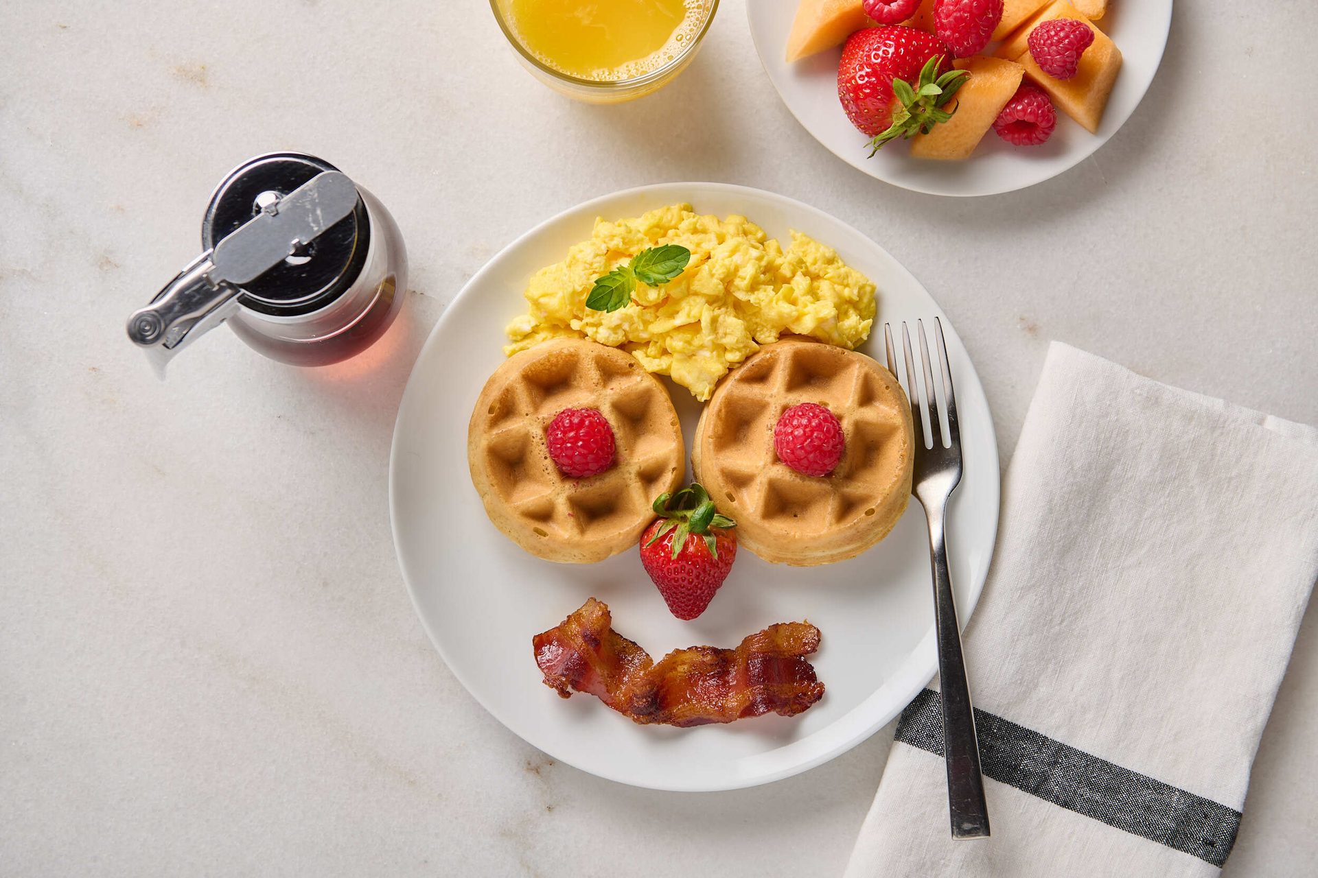 A cheerful breakfast plate featuring a smiley face made of waffles, scrambled eggs, strawberry, and bacon. Served with fruit, juice, and syrup.