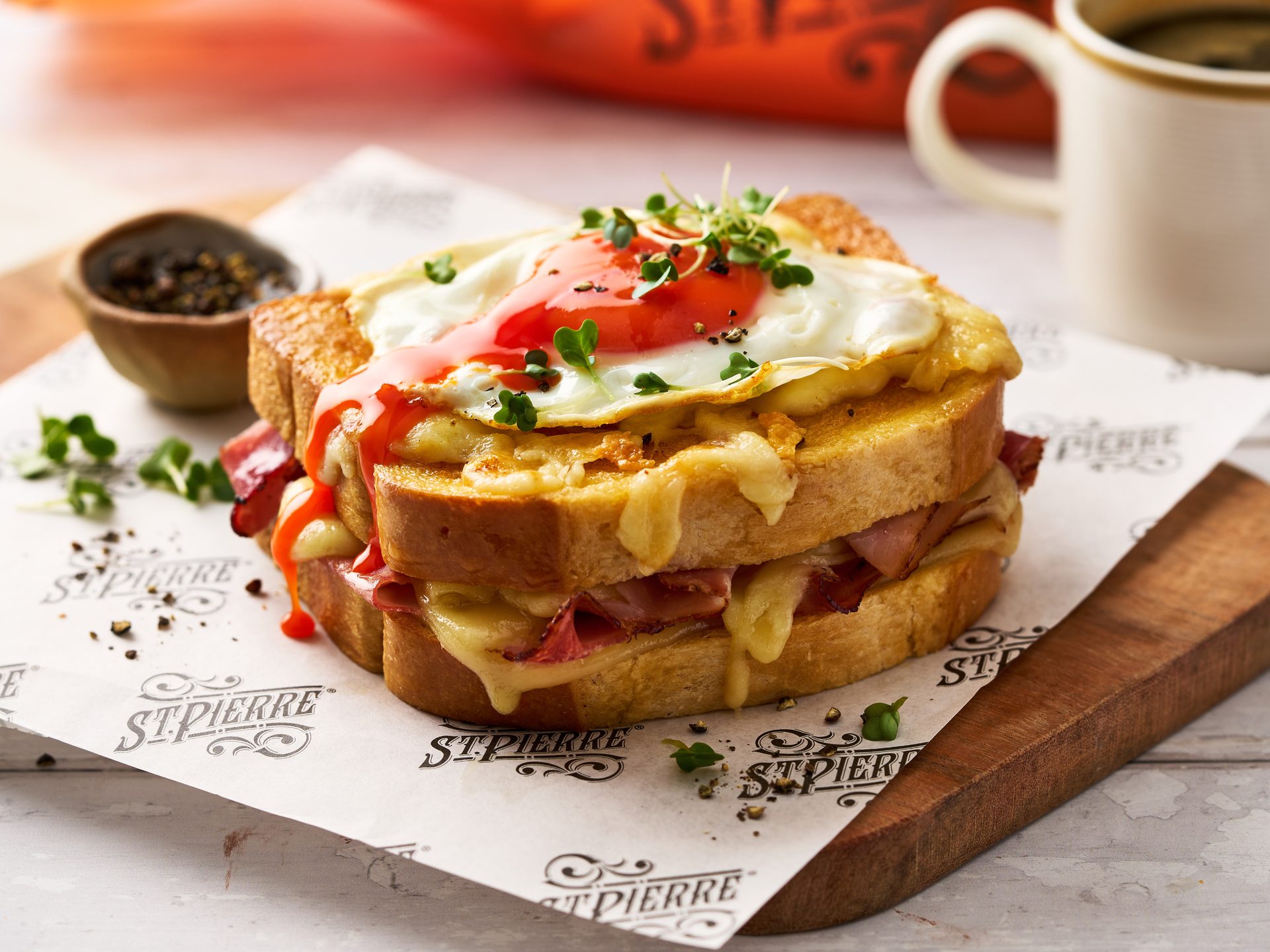 A cheesy croque madame sandwich topped with a fried egg, red sauce, and microgreens on branded paper.