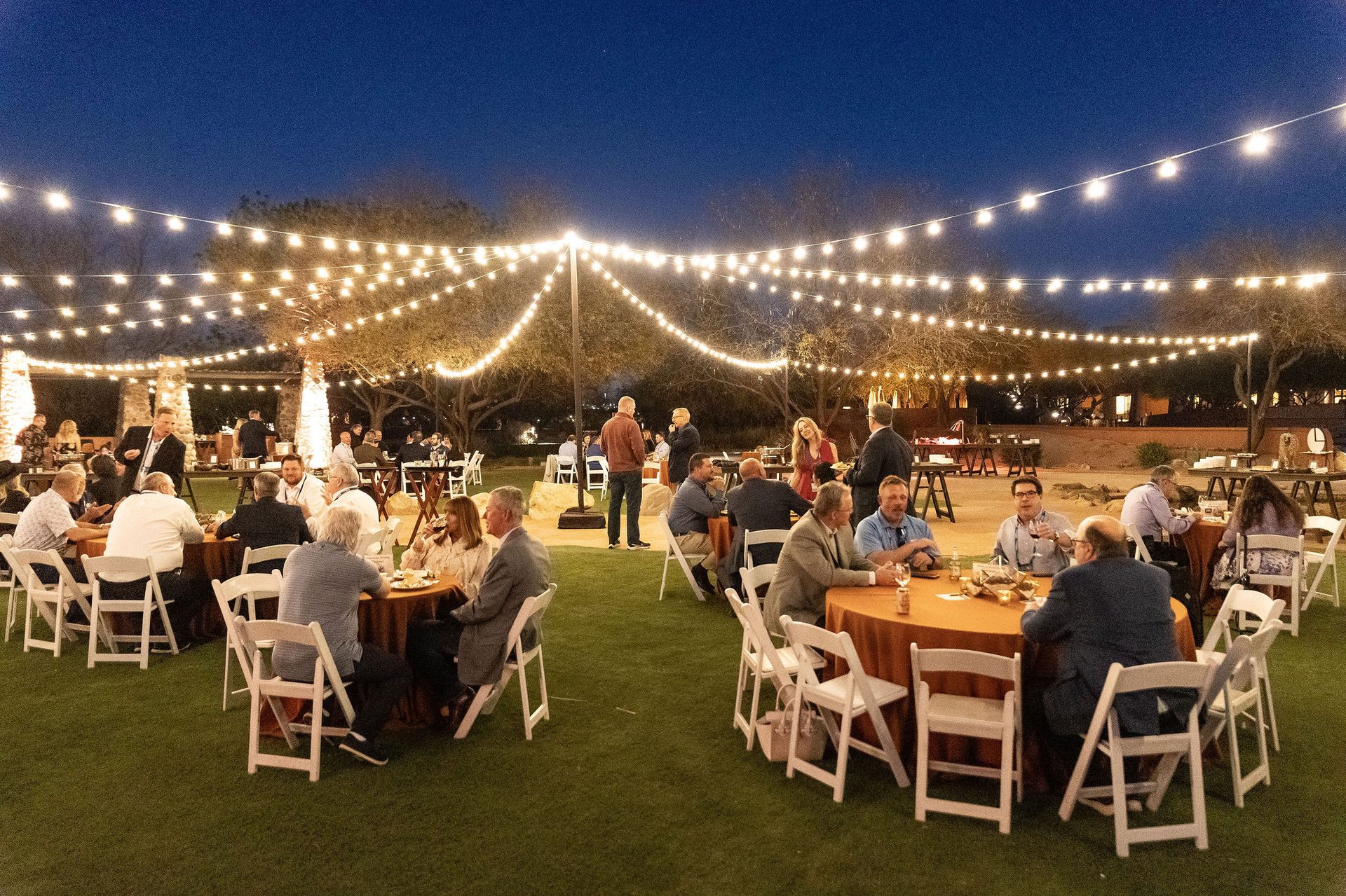 Outdoor evening event with people dining at tables under string lights.