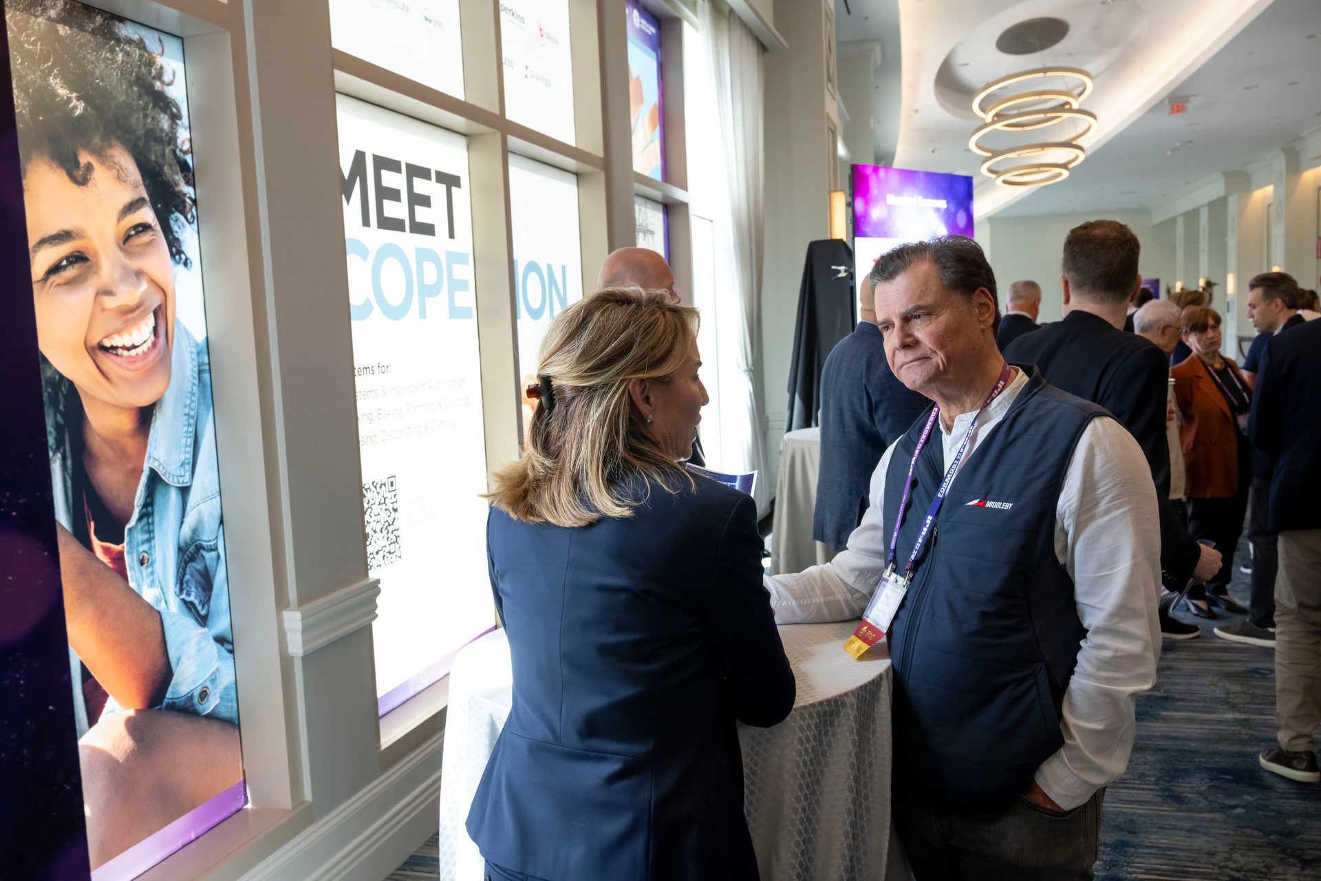 Two attendees converse at a networking event, with a large smiling woman poster visible on the left.