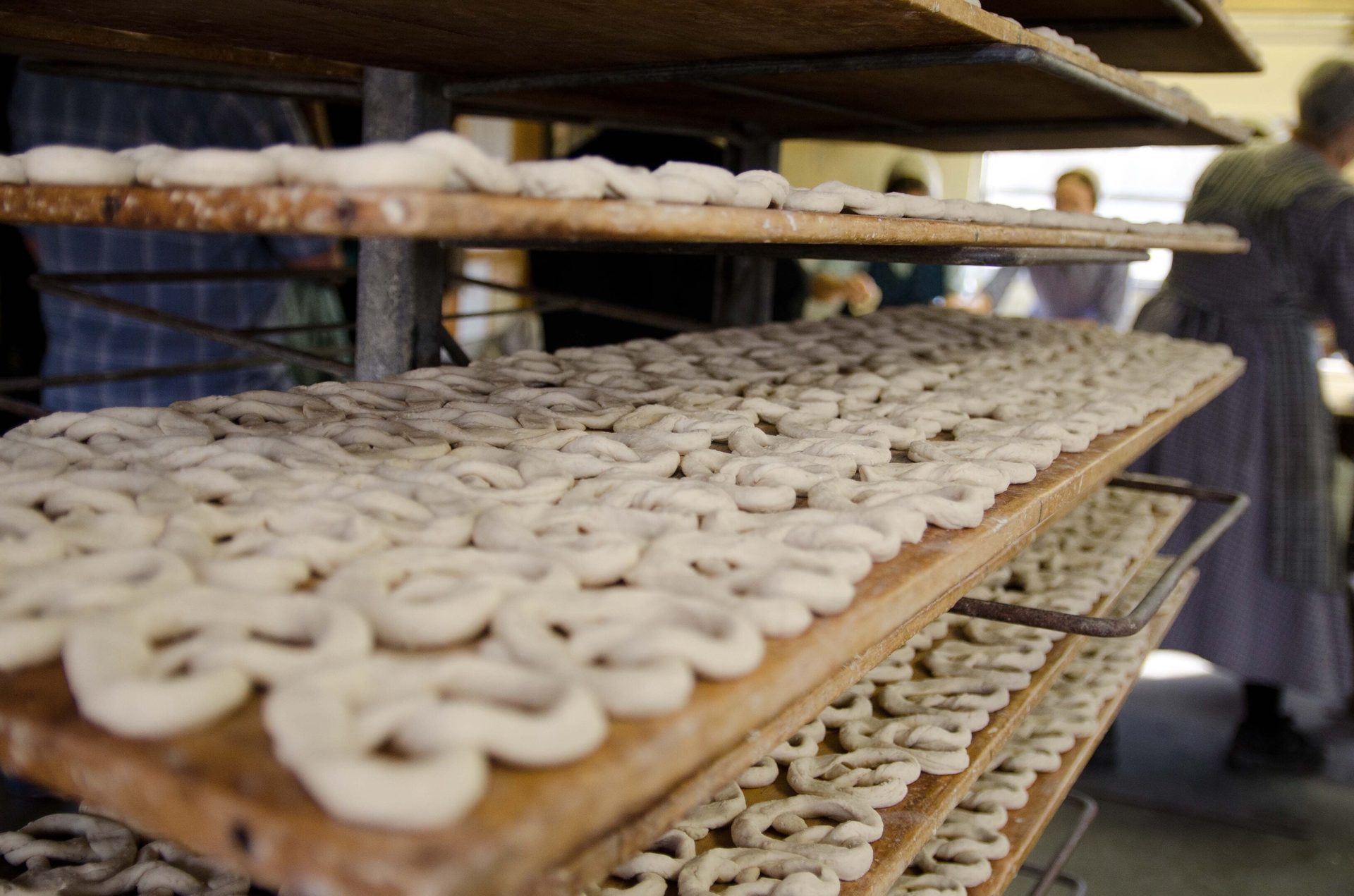 Rows of unbaked pretzels on wooden racks with bakers working in the blurry background.