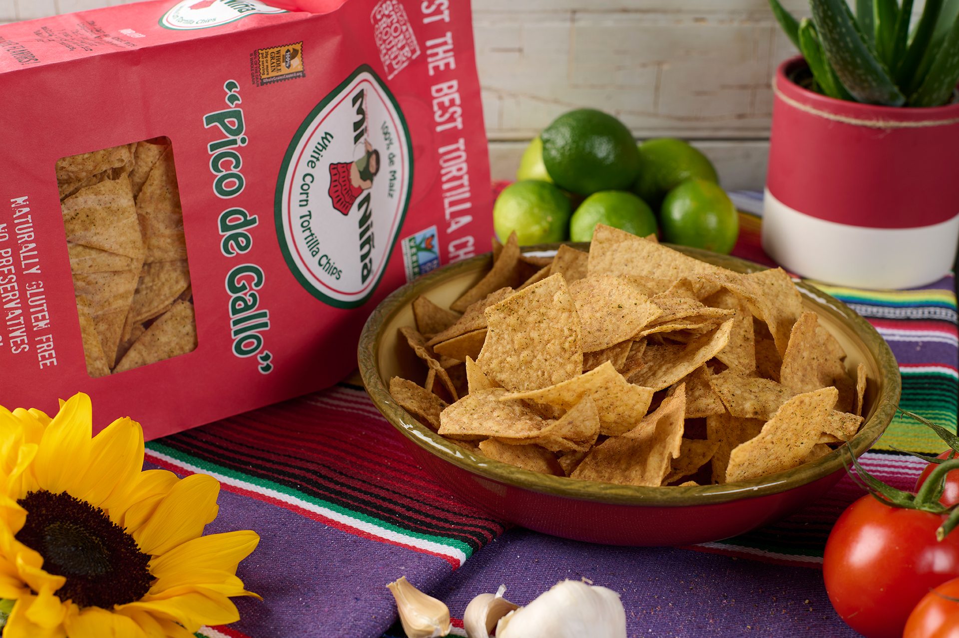 Mi Niña Pico de Gallo tortilla chips bag and bowl, with limes, tomatoes, garlic, and a sunflower.