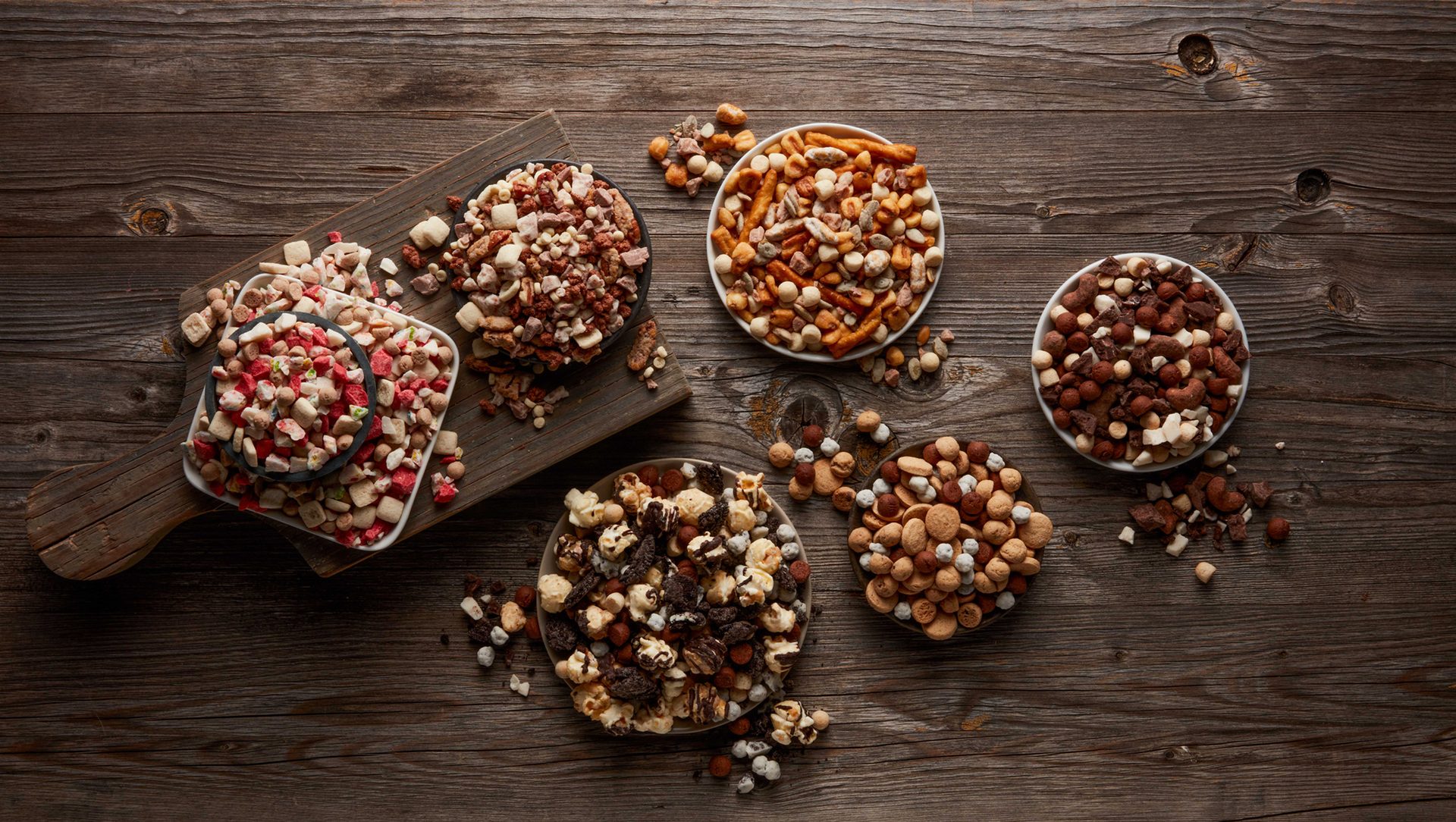 Bowls of assorted mixed snacks (cereals, dried fruits, nuts, pretzels, popcorn) on a rustic wooden table.