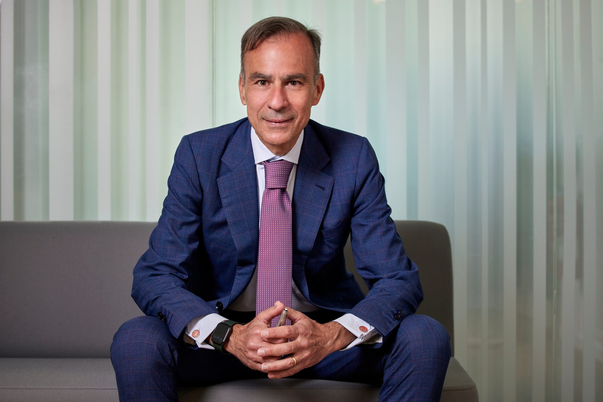 A man in a blue checkered suit and purple tie, sitting on a gray couch, smiling at the camera.