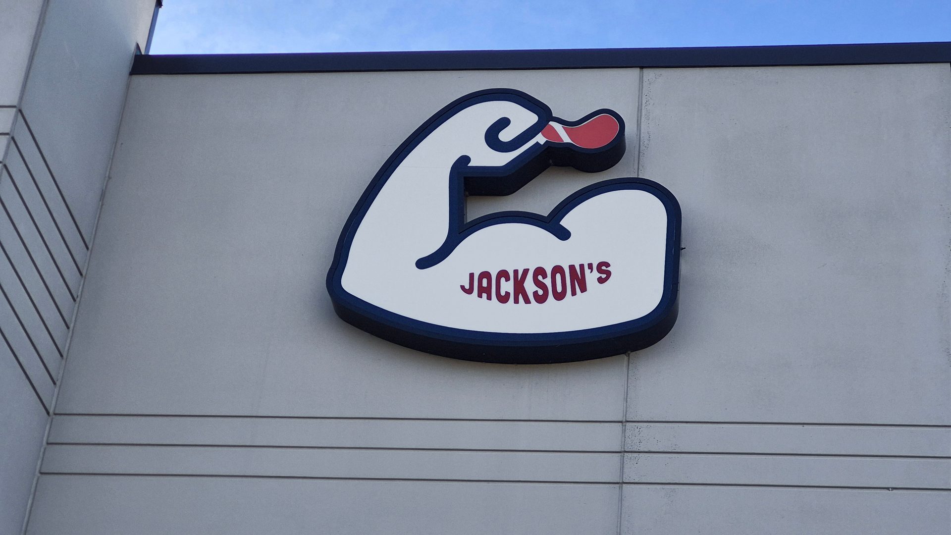A white, blue, and red sign shaped like a flexing arm, with 'JACKSON'S' written on it, mounted on a building.