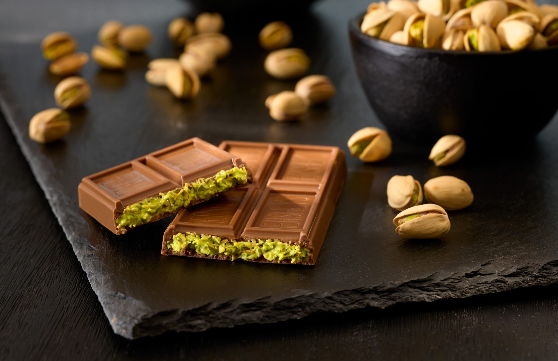 Pistachio-filled chocolate bar, loose pistachios, and a bowl of pistachios on dark slate.
