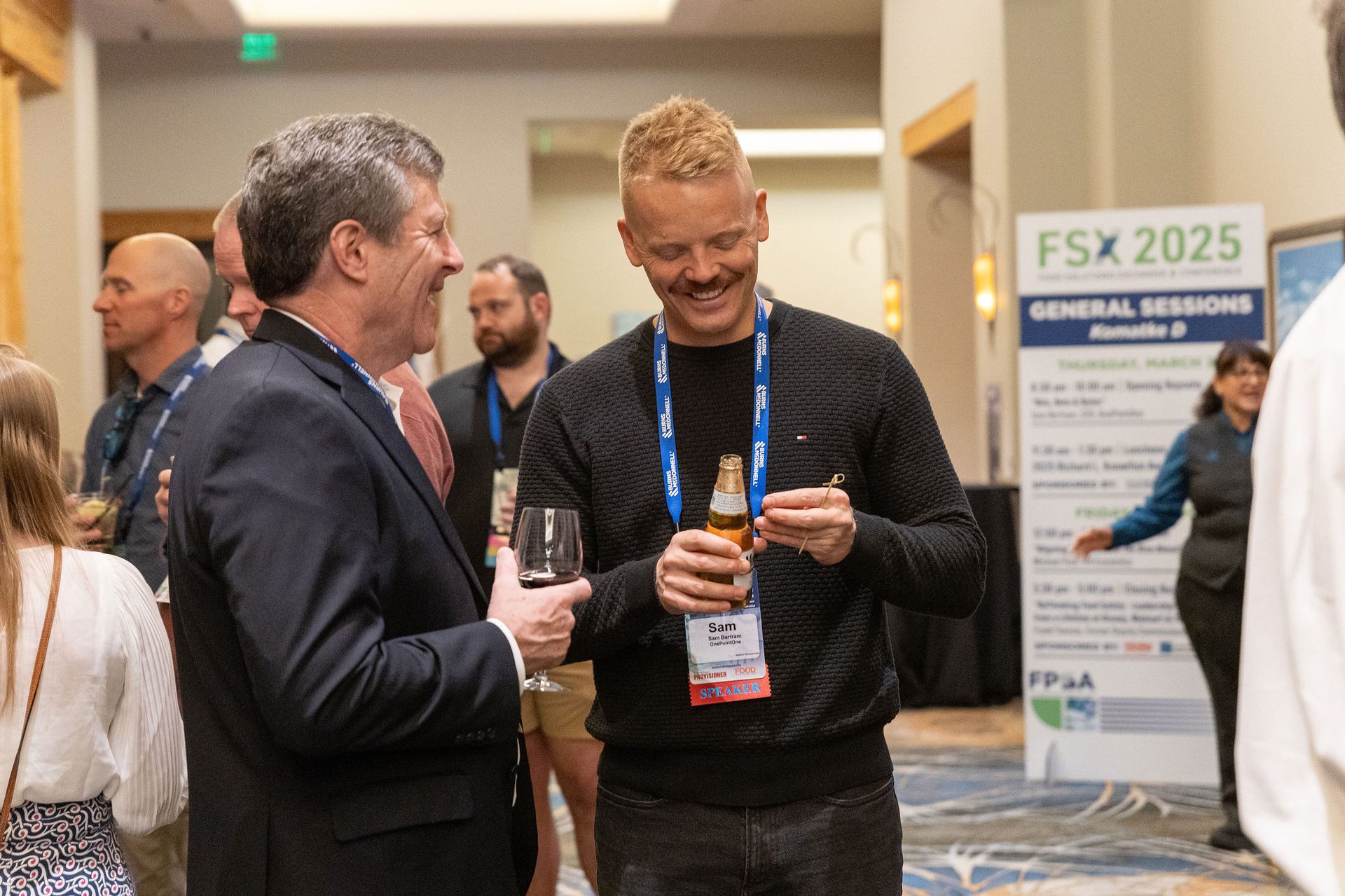 Two men smiling and talking at a conference, holding drinks, with an "FSX 2025" sign.