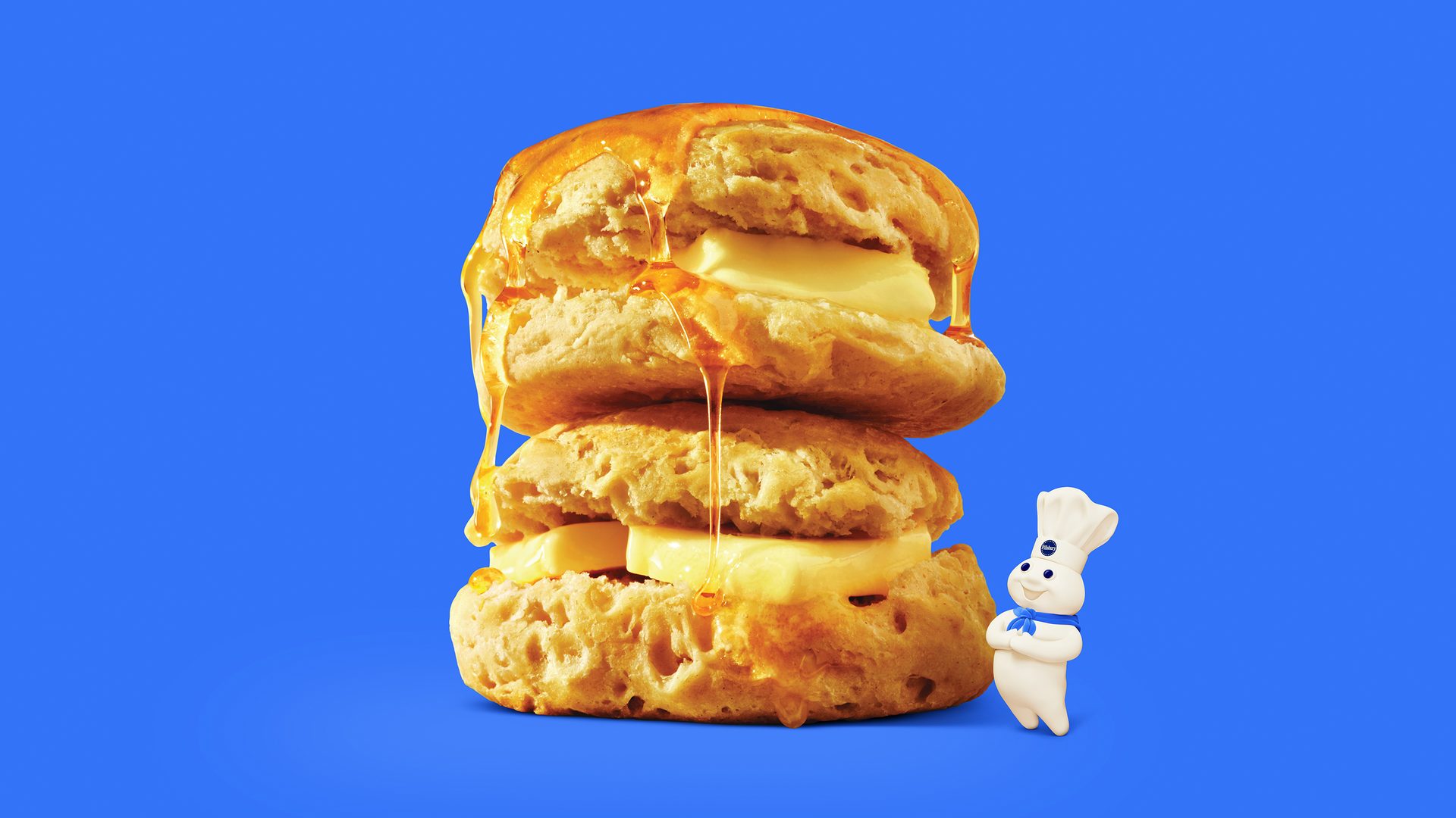 Pillsbury Doughboy next to honey-drenched stacked biscuits with butter on a blue background.