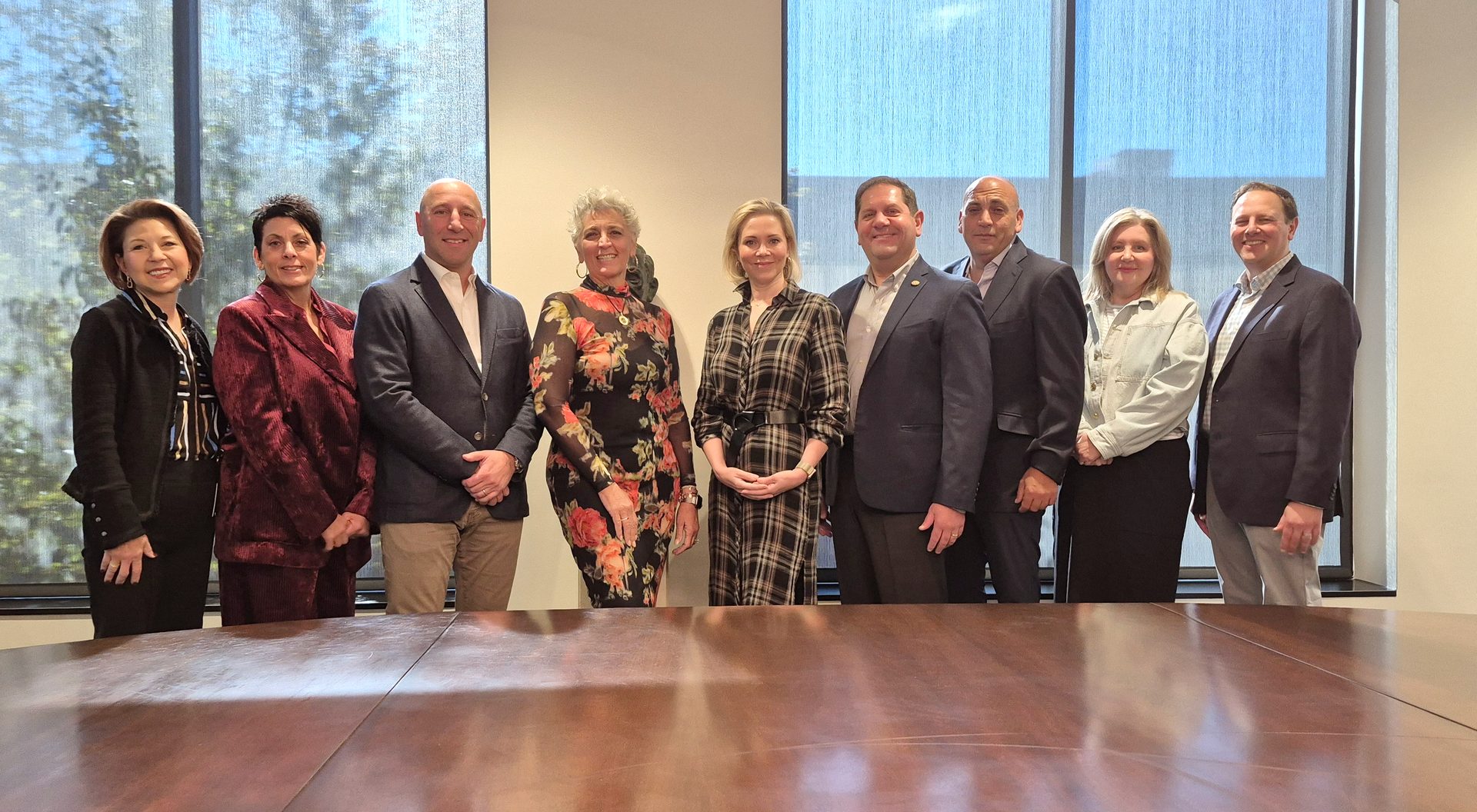 Nine professionals (four women, five men) smiling, standing in a bright room with large windows.