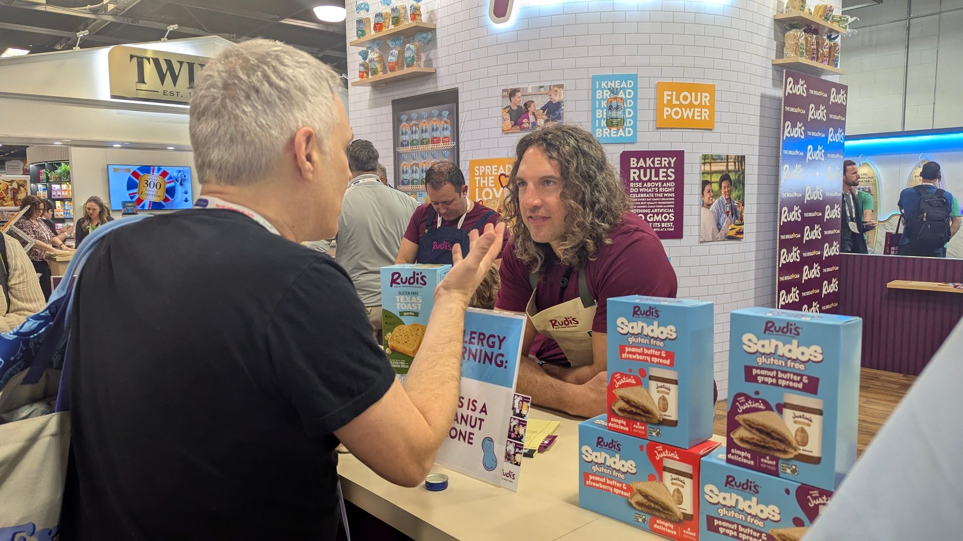 Two men converse at a Rudi's gluten-free food stand, showcasing Sandos products.
