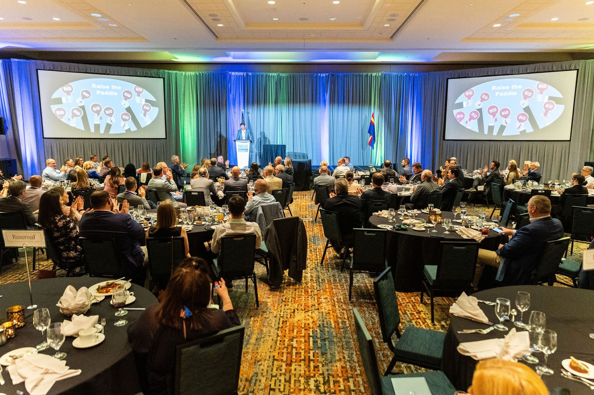 Ballroom event: speaker, audience at tables, screens display "Raise the Paddle BID."
