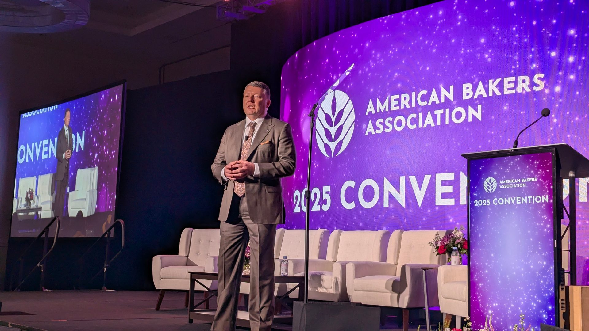Man speaking at American Bakers Association 2025 Convention with large screens and a podium.