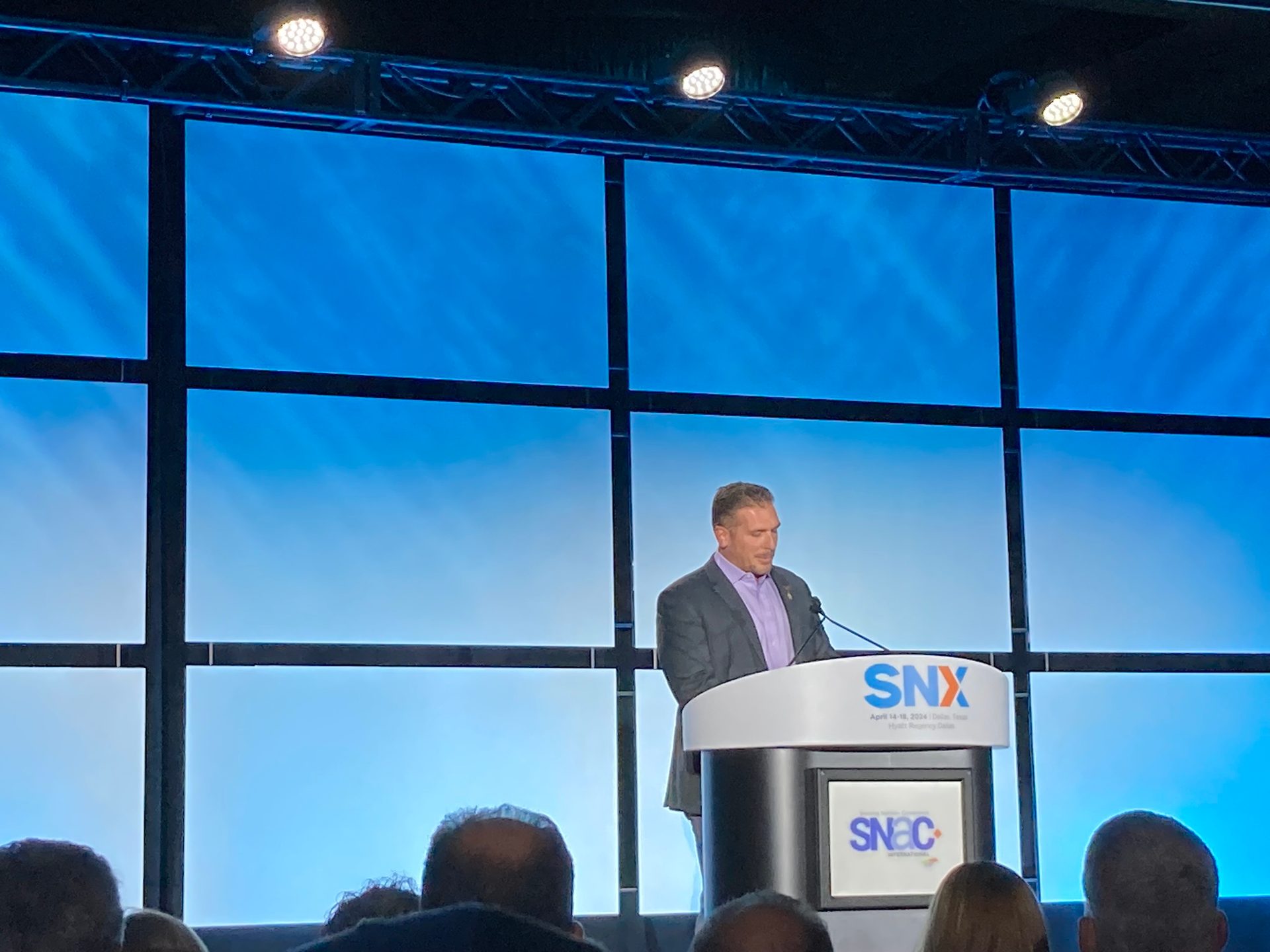Man speaking at podium with "SNX" logo, "SNAC" on screen, blue background.