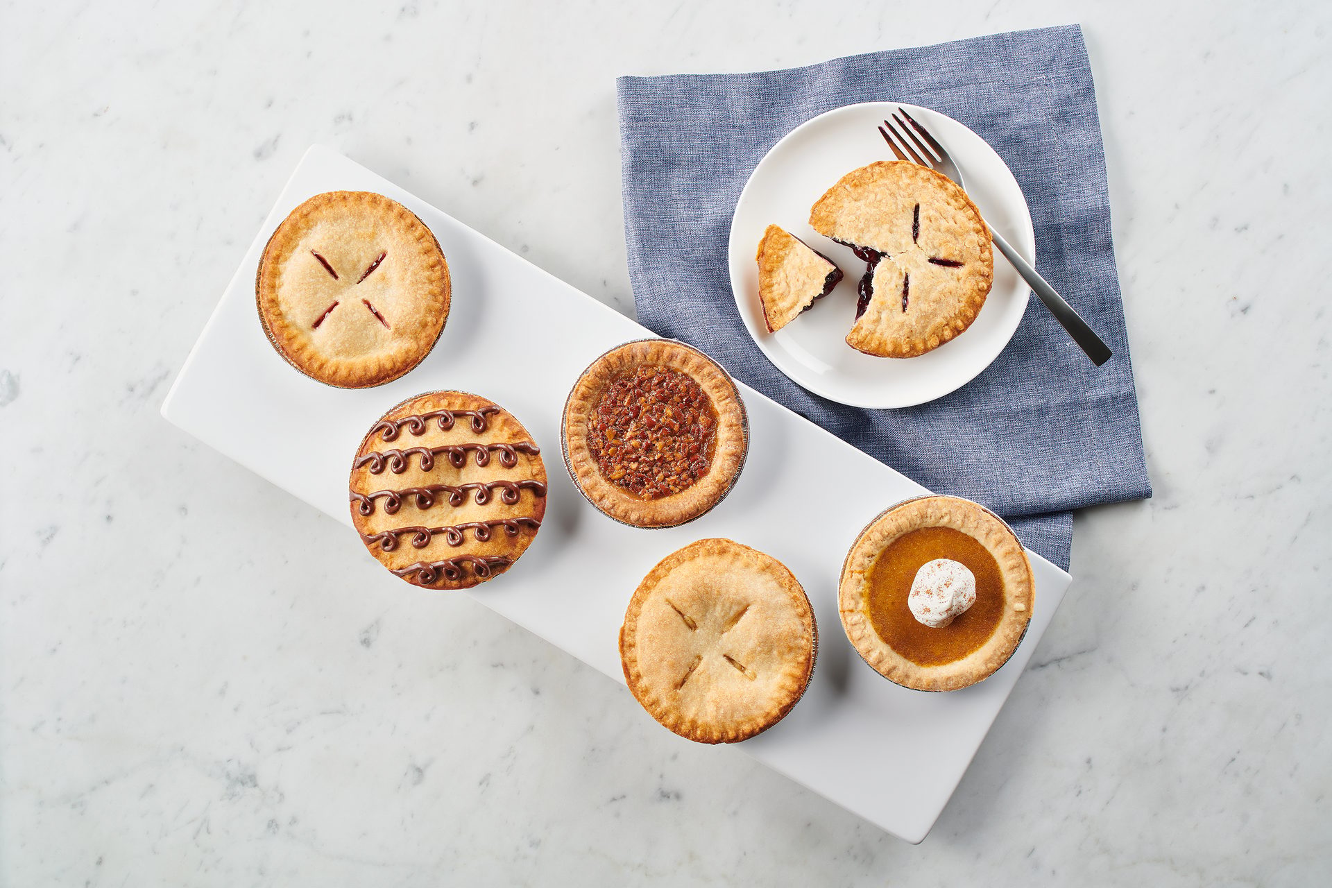 Assorted mini pies, including berry, pecan, pumpkin, and chocolate, on a white platter and plate.