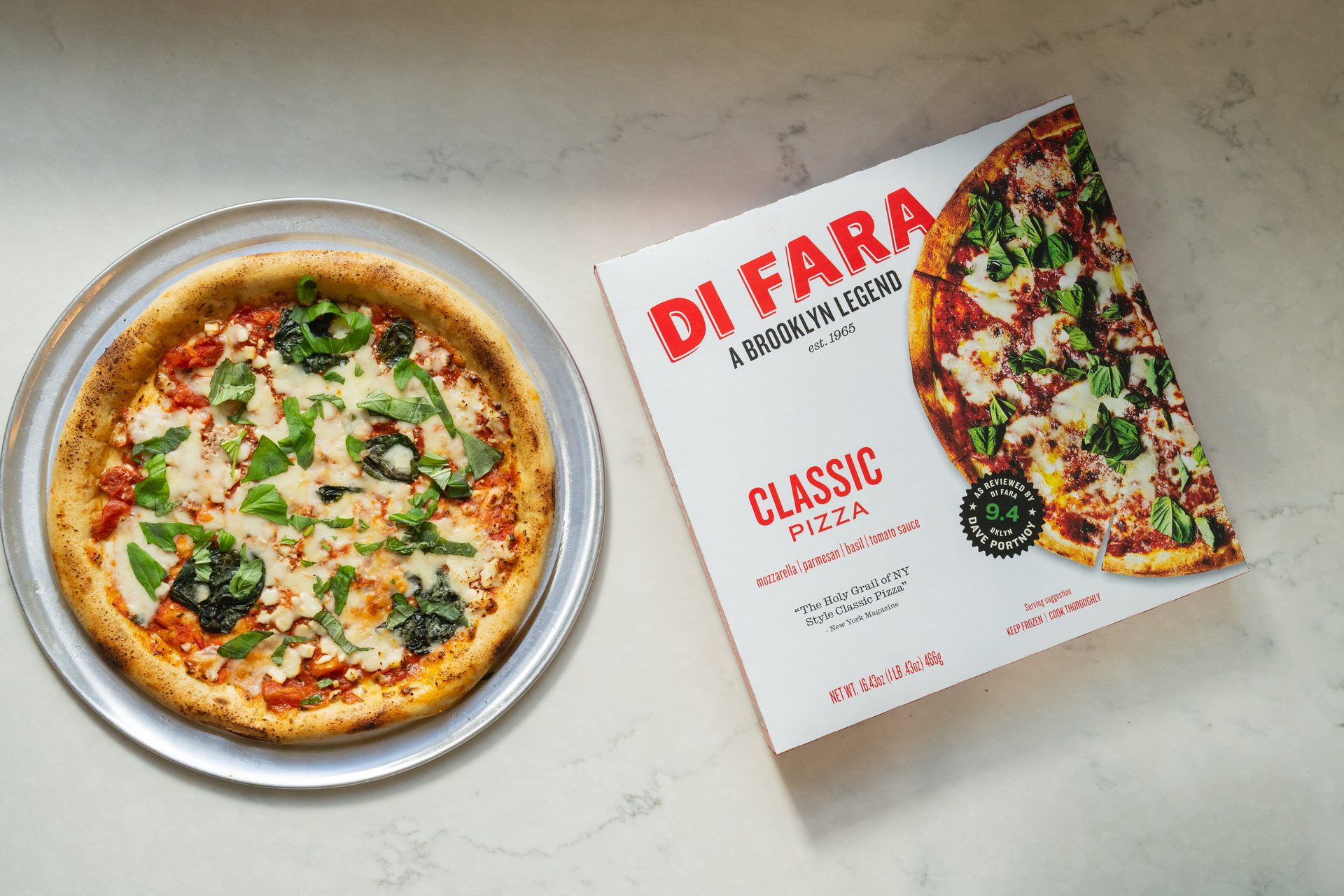 A freshly baked Di Fara Classic Pizza with basil and cheese next to its product box on a marble surface.