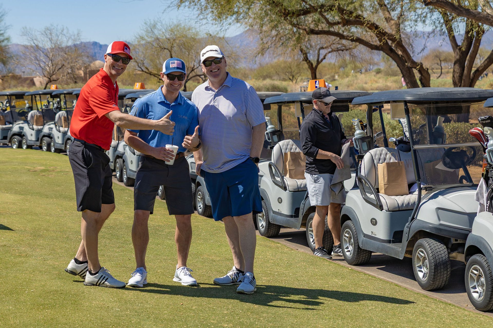 Three golfers by carts on a sunny course, two smiling with thumbs up.