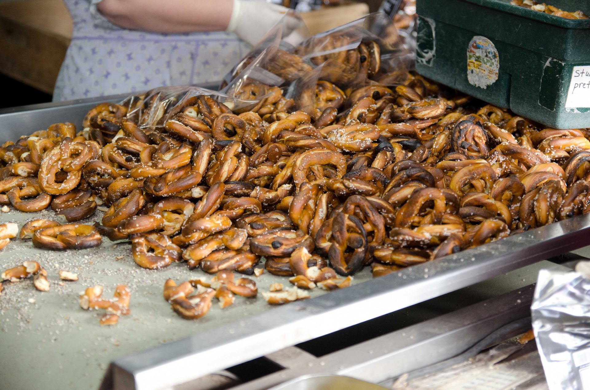 A large pile of dark brown, salted pretzels on a metal tray, with a person and container in the background.