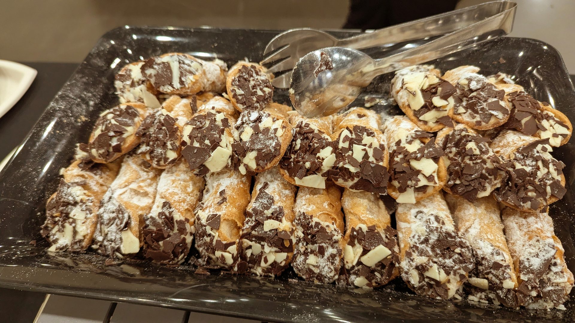 A black platter piled with cannoli, dusted with powdered sugar and topped with dark and white chocolate shavings.