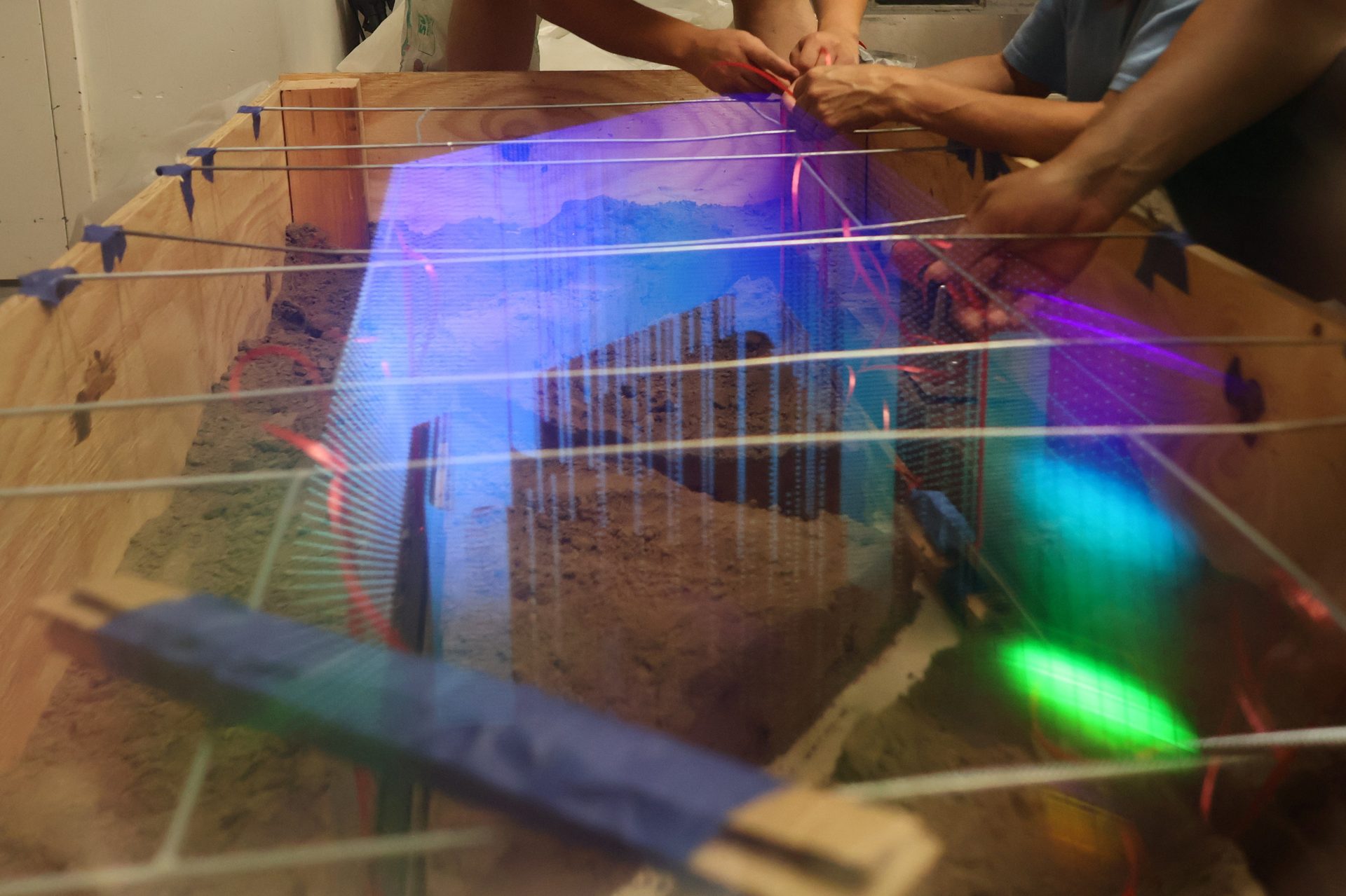 Hands adjusting wires over sand in a wooden box, with blue, purple, and green light.