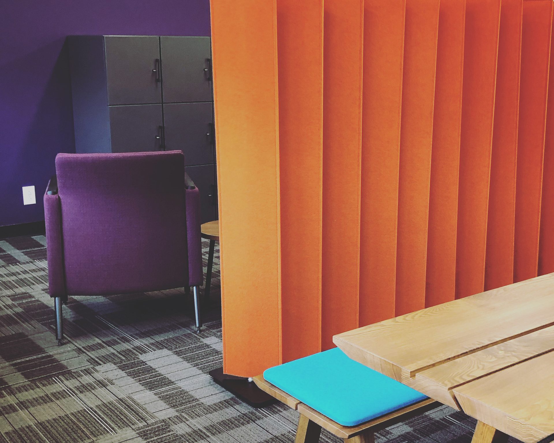 A colorful office space with a purple chair, orange divider, wooden table, and blue cushion.