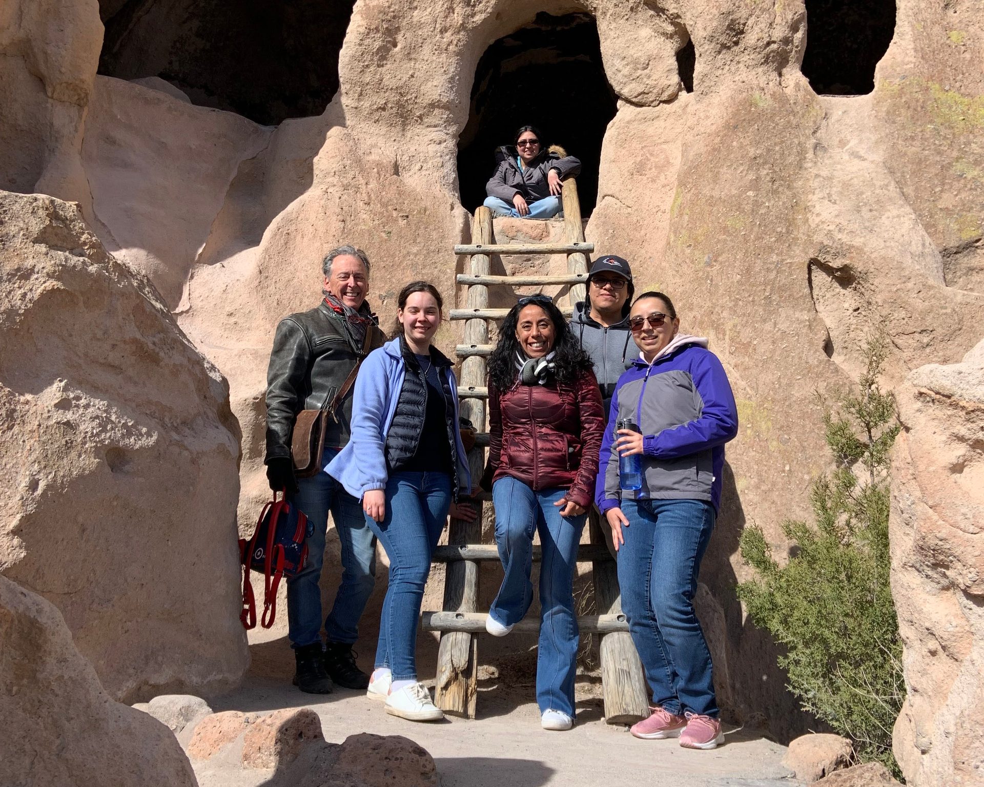 Six people, some standing, one on a ladder, pose in front of cave dwellings carved into rock formations.
