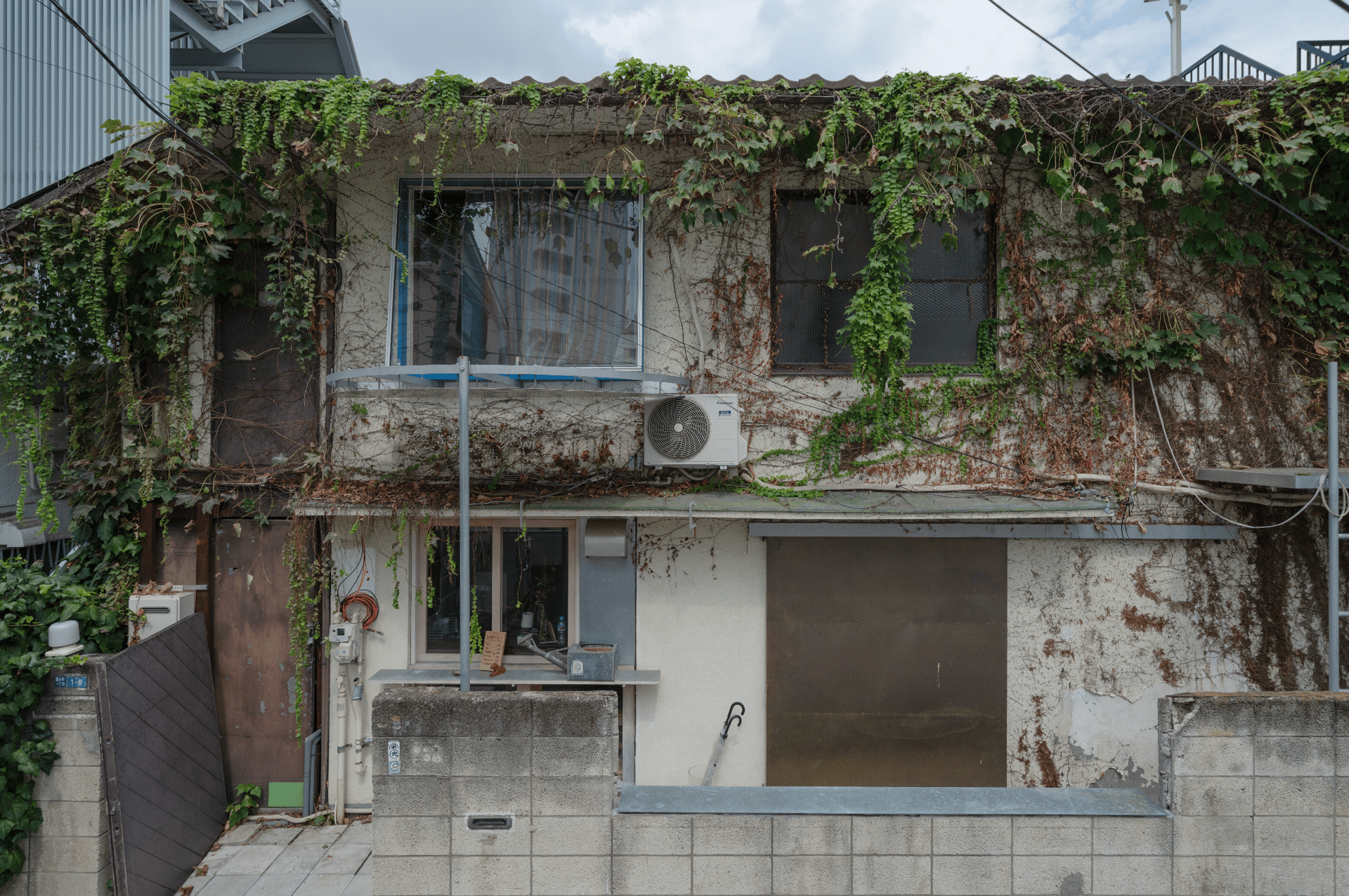 An old, ivy-covered building with windows, an AC unit, and a concrete wall at its base.