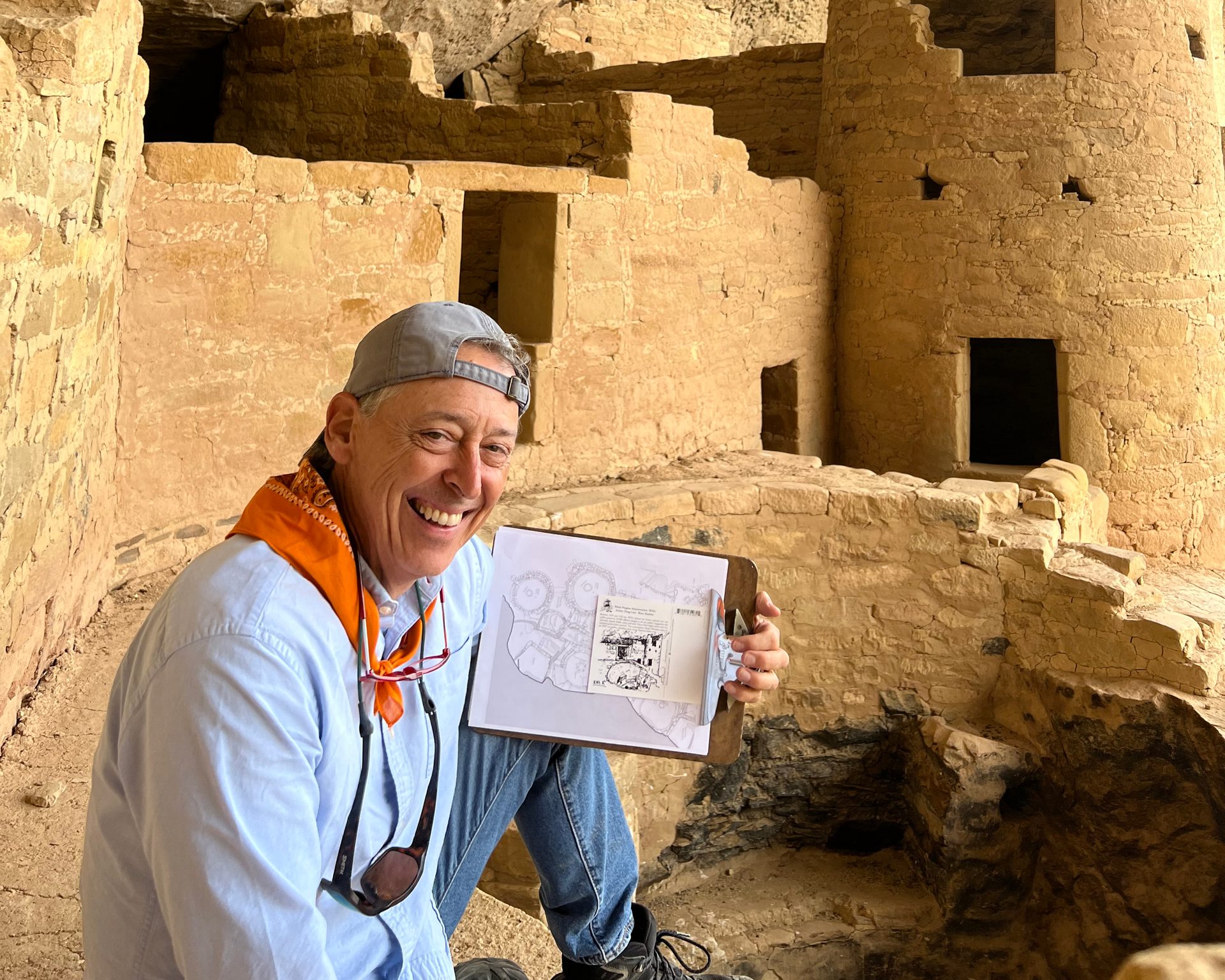 Smiling man holds a map on a clipboard in front of ancient cliff dwellings.