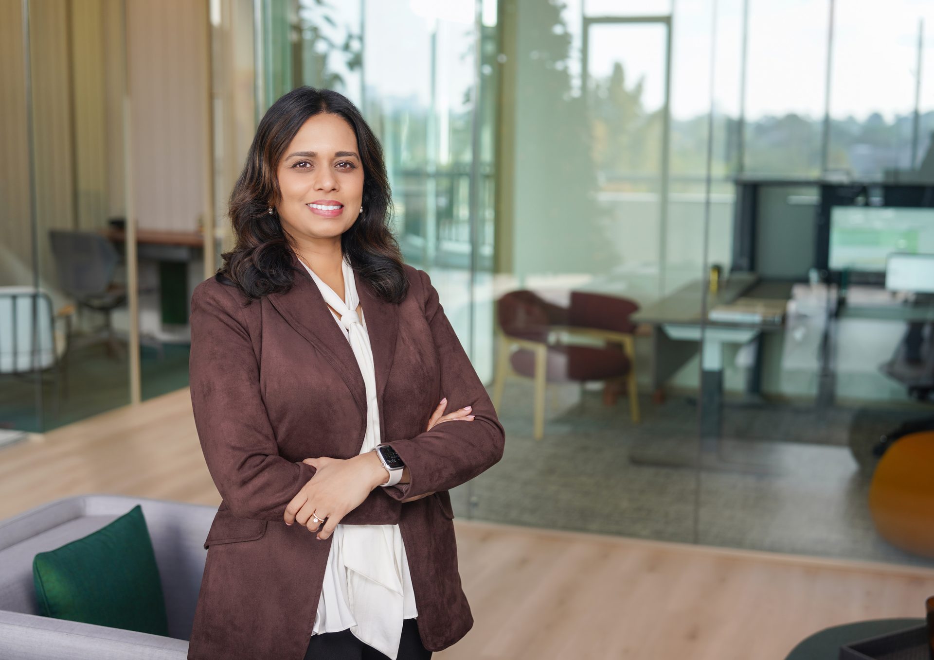 A professional woman in a brown blazer and white blouse smiles, standing with crossed arms in an office.