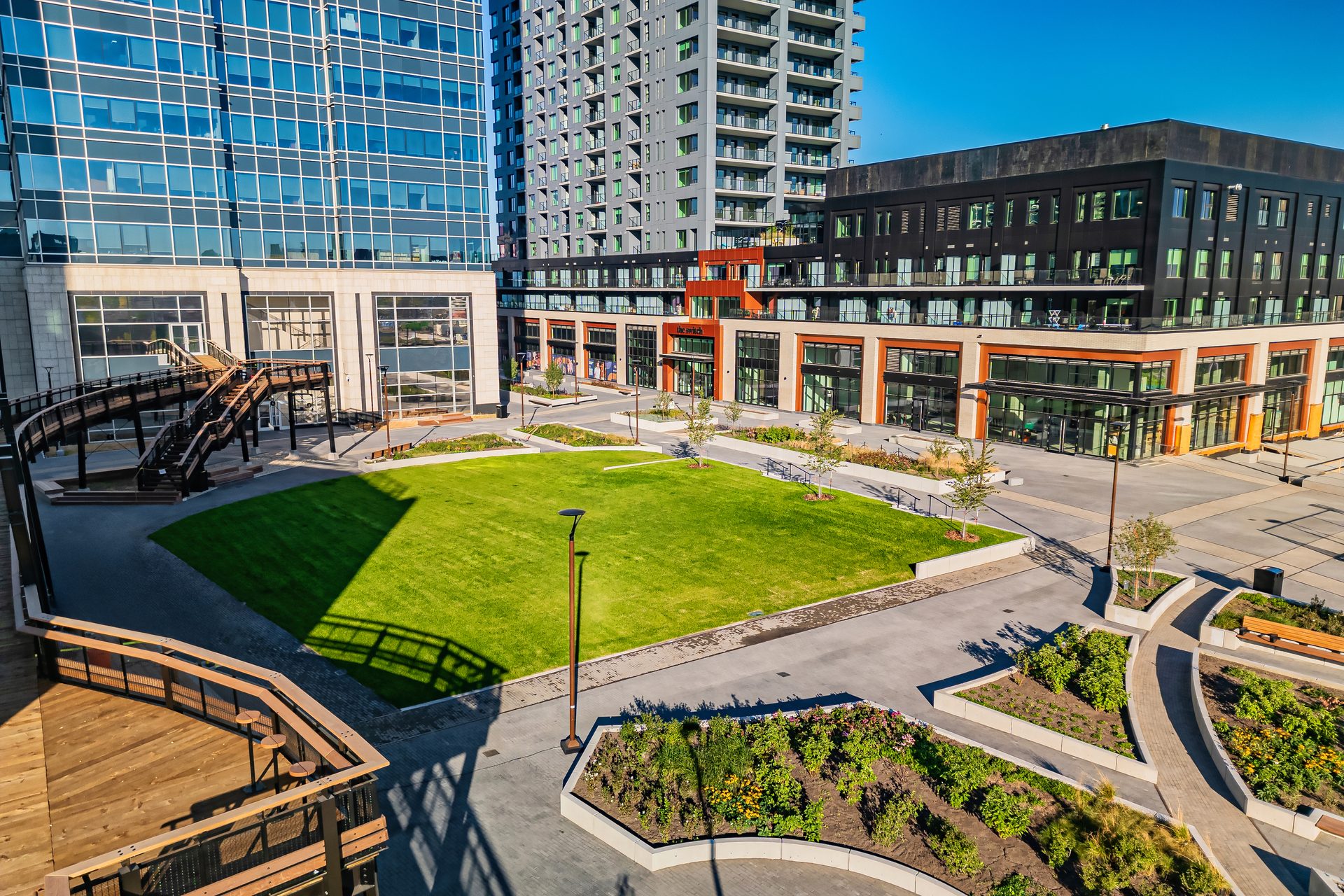 Overhead view: Modern city plaza with glass buildings, green lawn, and elevated wooden path.