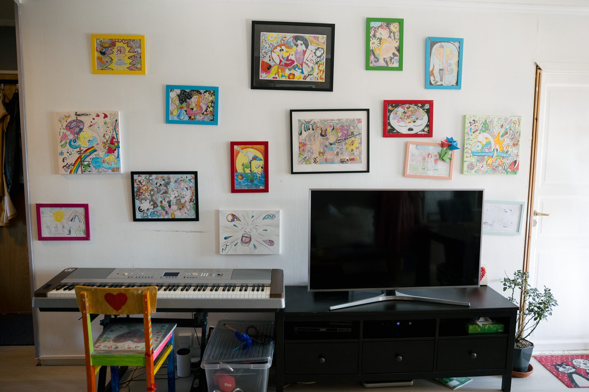 en hvit vegg i en leilighet med en rekke fargerike bilder innrammet i ulike farger. I forgrunnen er det et tv-bord, en TV, en farget stol og et gr&#xE5;tt keyboard.