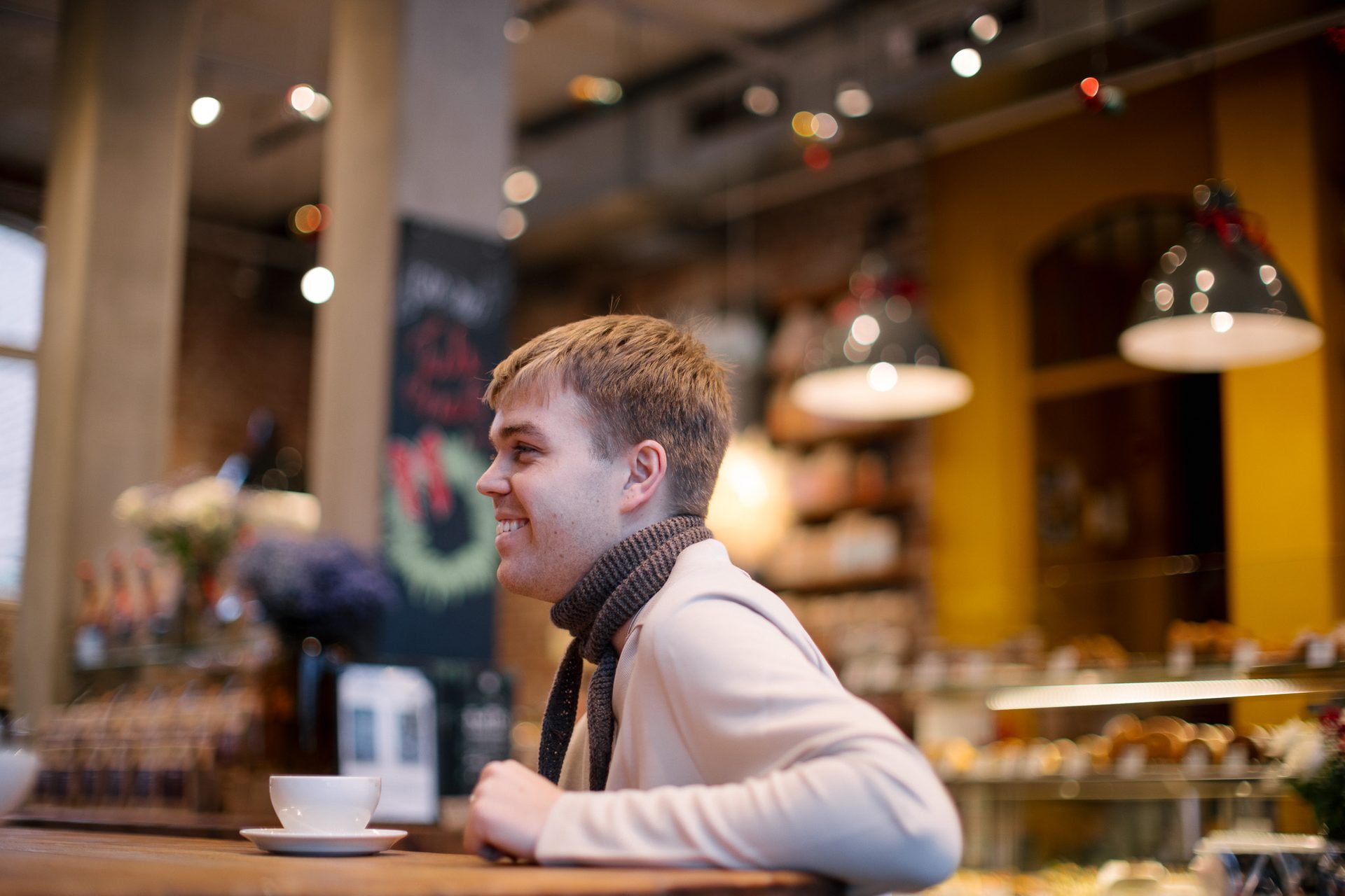 En mann i beige poloskjorte og skjerf sitter med en kaffekopp i en kaf&#xE9;. Han ser bort mot noen og ler. 