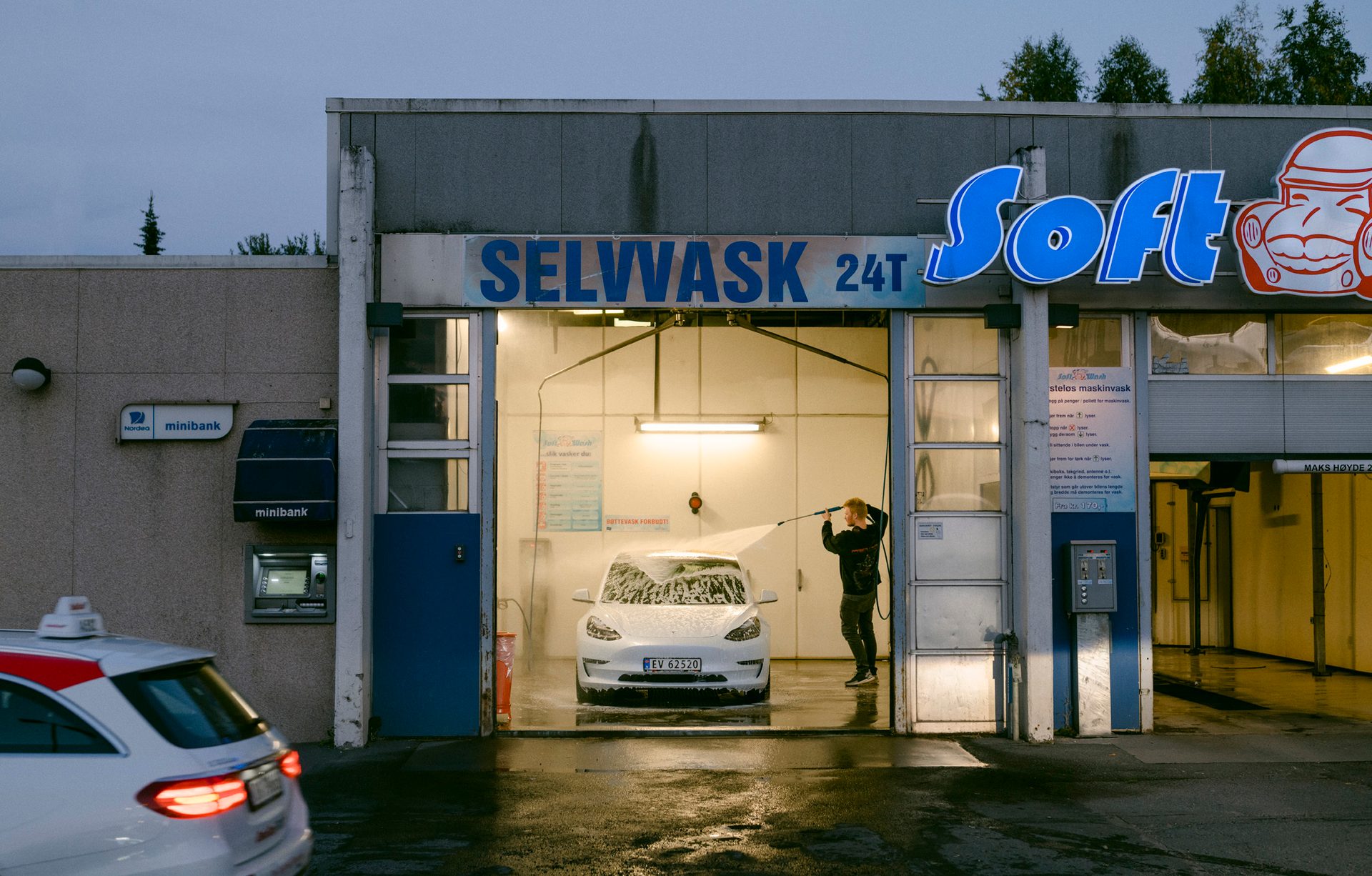 En ung mann spyler bilen sin i en vaskehall med selvvask p&#xE5; kvelden.