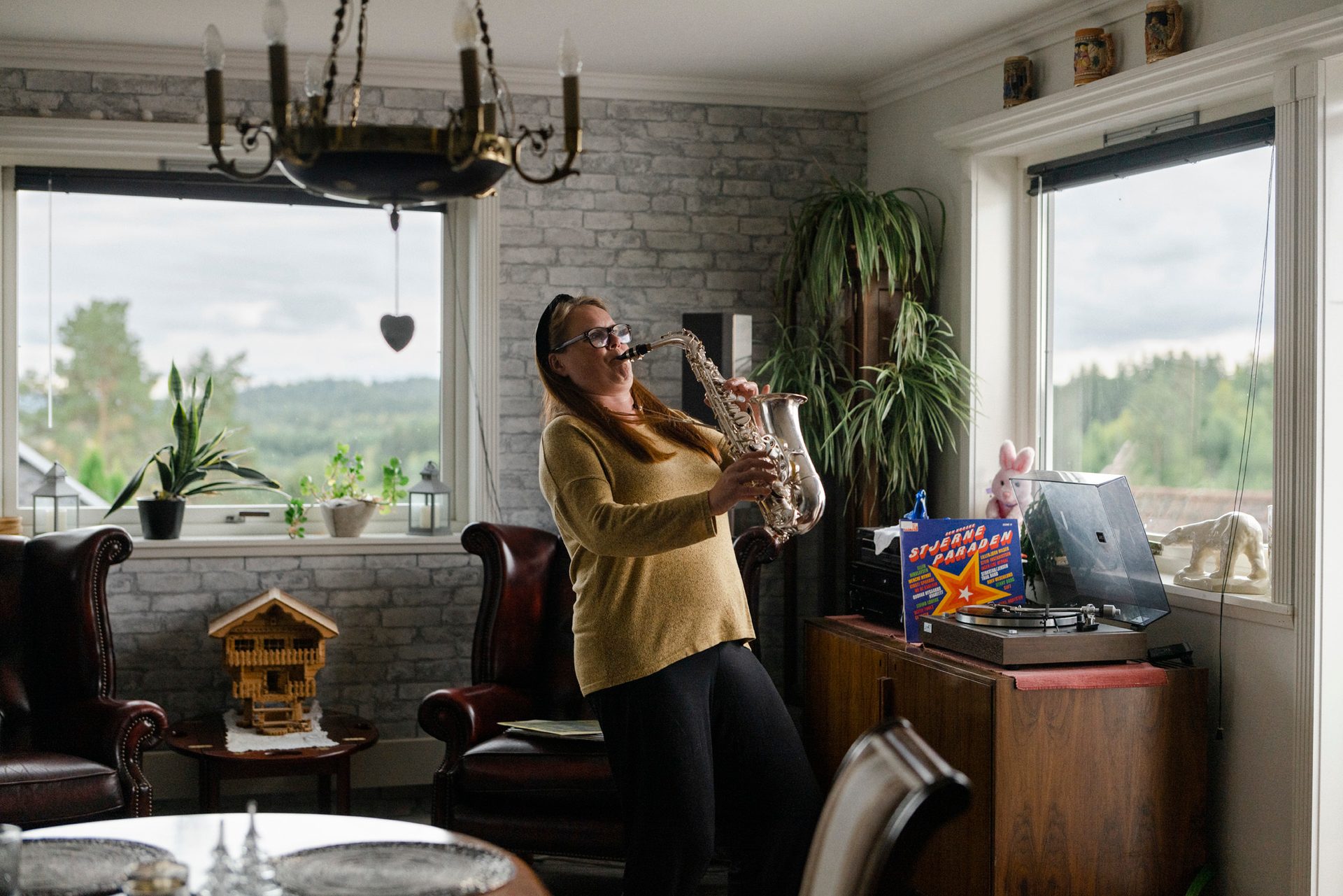 Kvinne spiller saksofon mens hun spiller LP-plate med musikk i stue med flere gr&#xF8;nne planter.