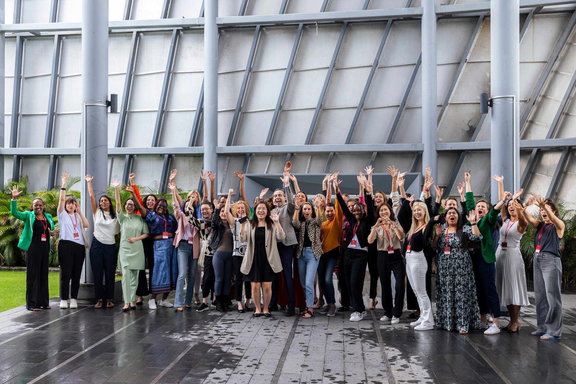 A large group of diverse people, mostly women, raising their hands in excitement and smiling brightly outdoors.