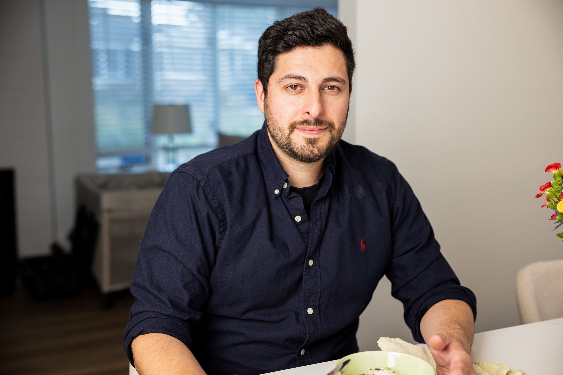 A smiling man with a beard and dark blue shirt sits at a table with flowers and food.