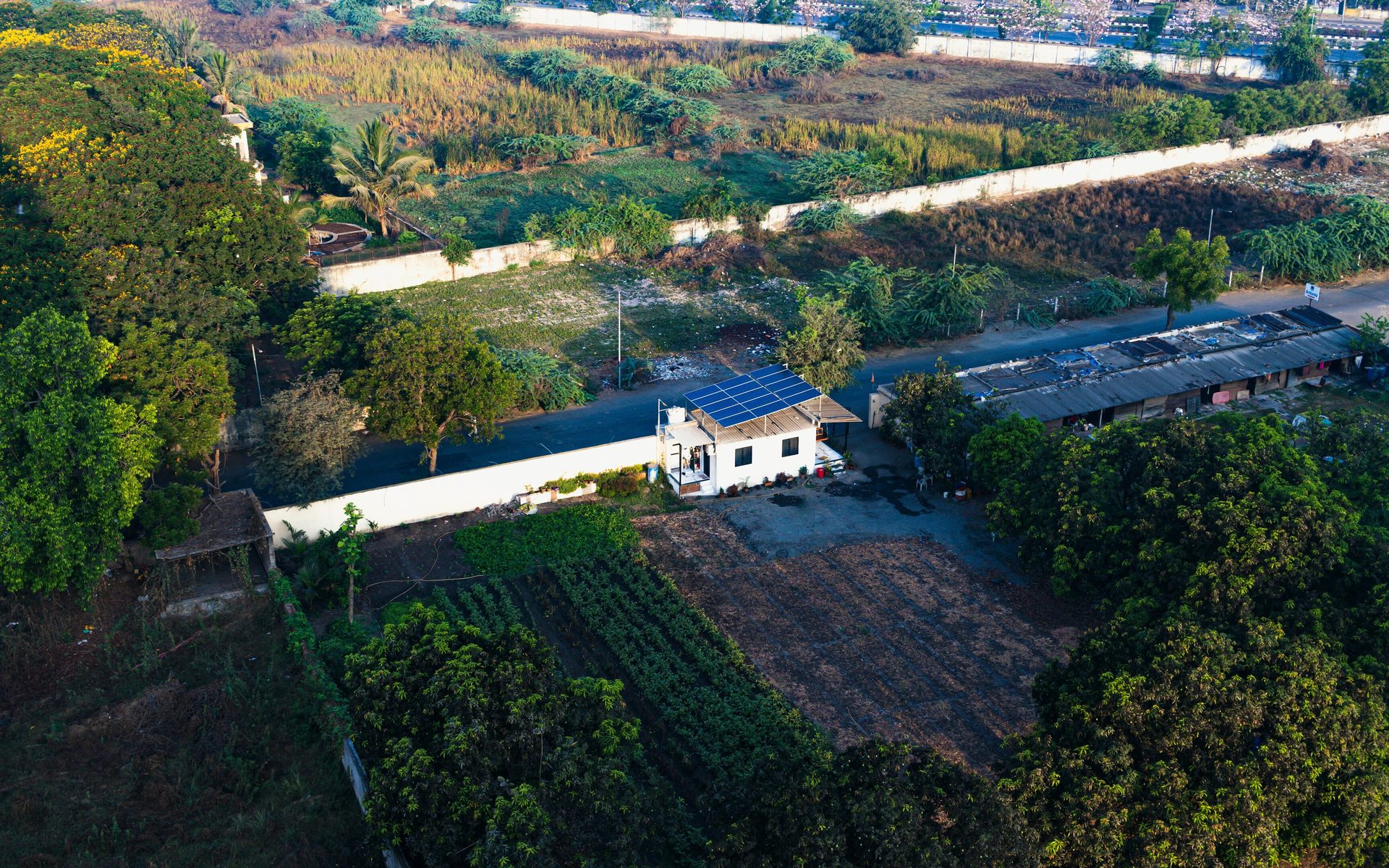 Aerial farm: house with solar panels, fields, trees, and long buildings.