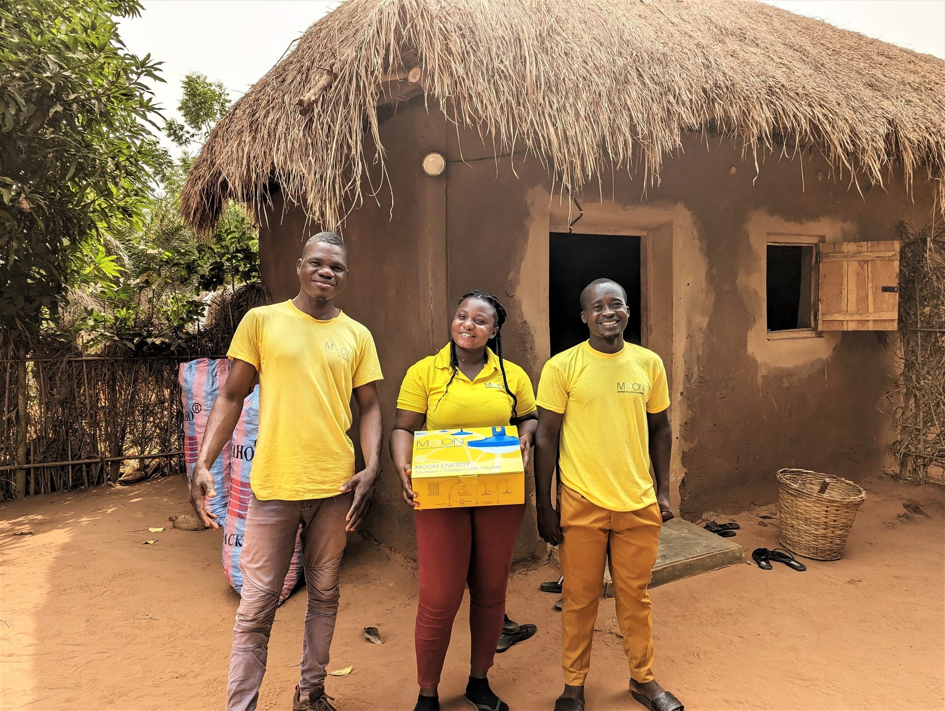 Three people in yellow Moon Energy shirts hold a box by a house.