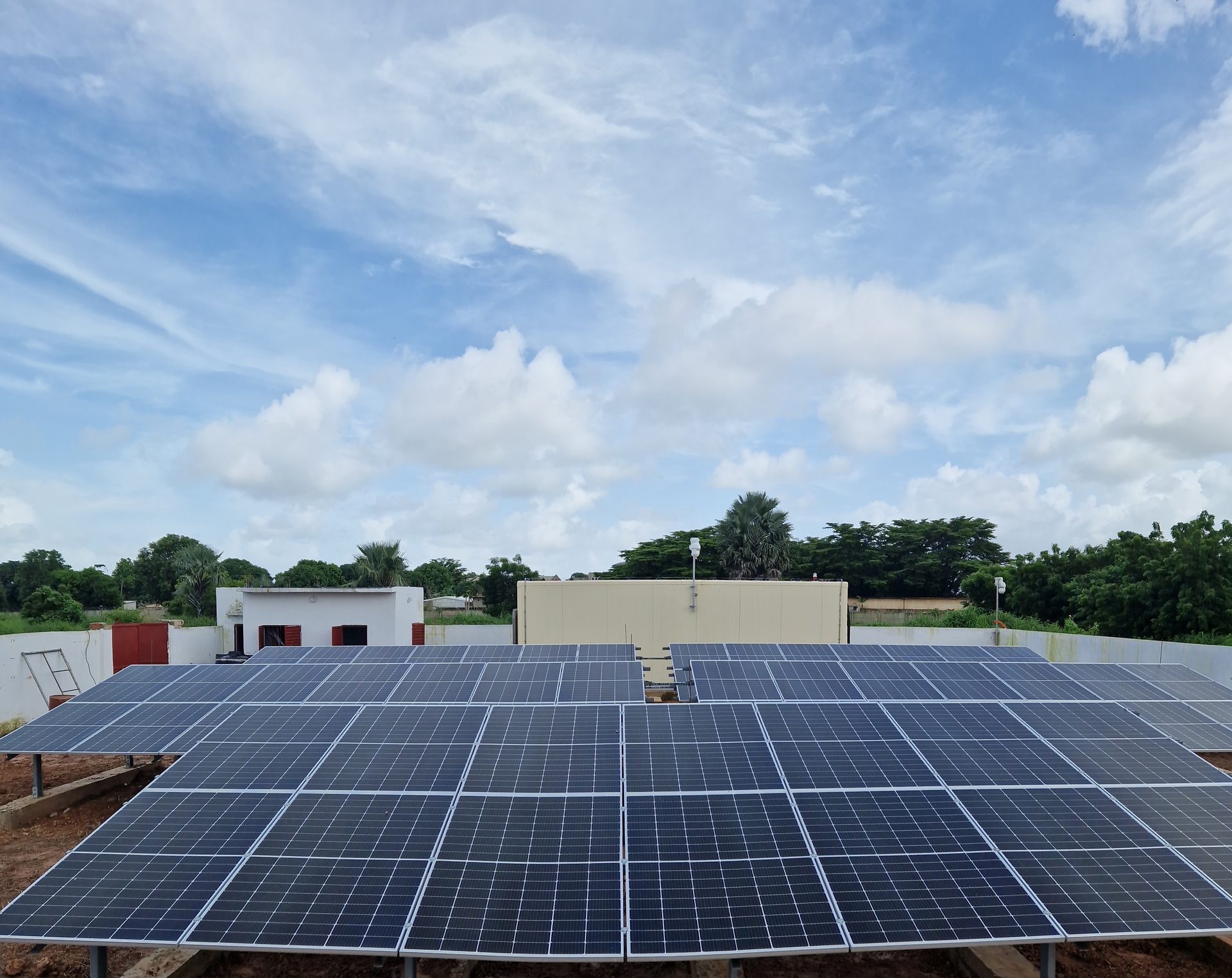 Solar panel array with buildings and trees under a cloudy sky.