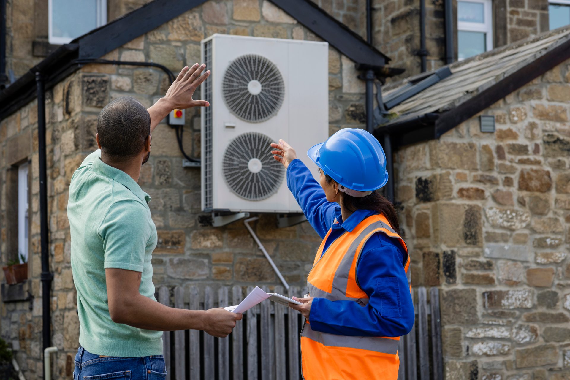 Technician and client pointing at an outdoor AC unit on a stone house.