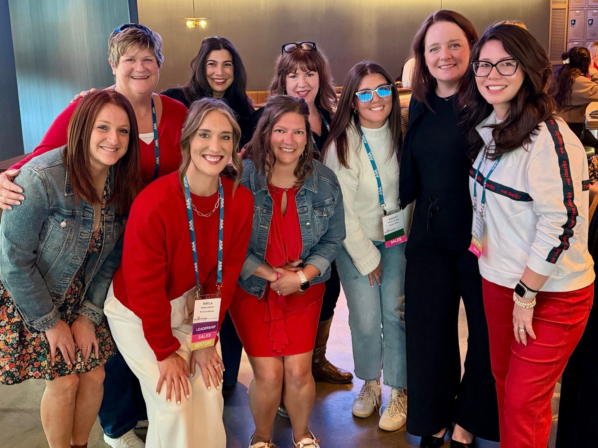 A group of nine smiling women posing together at an event, some wearing name tags and lanyards.