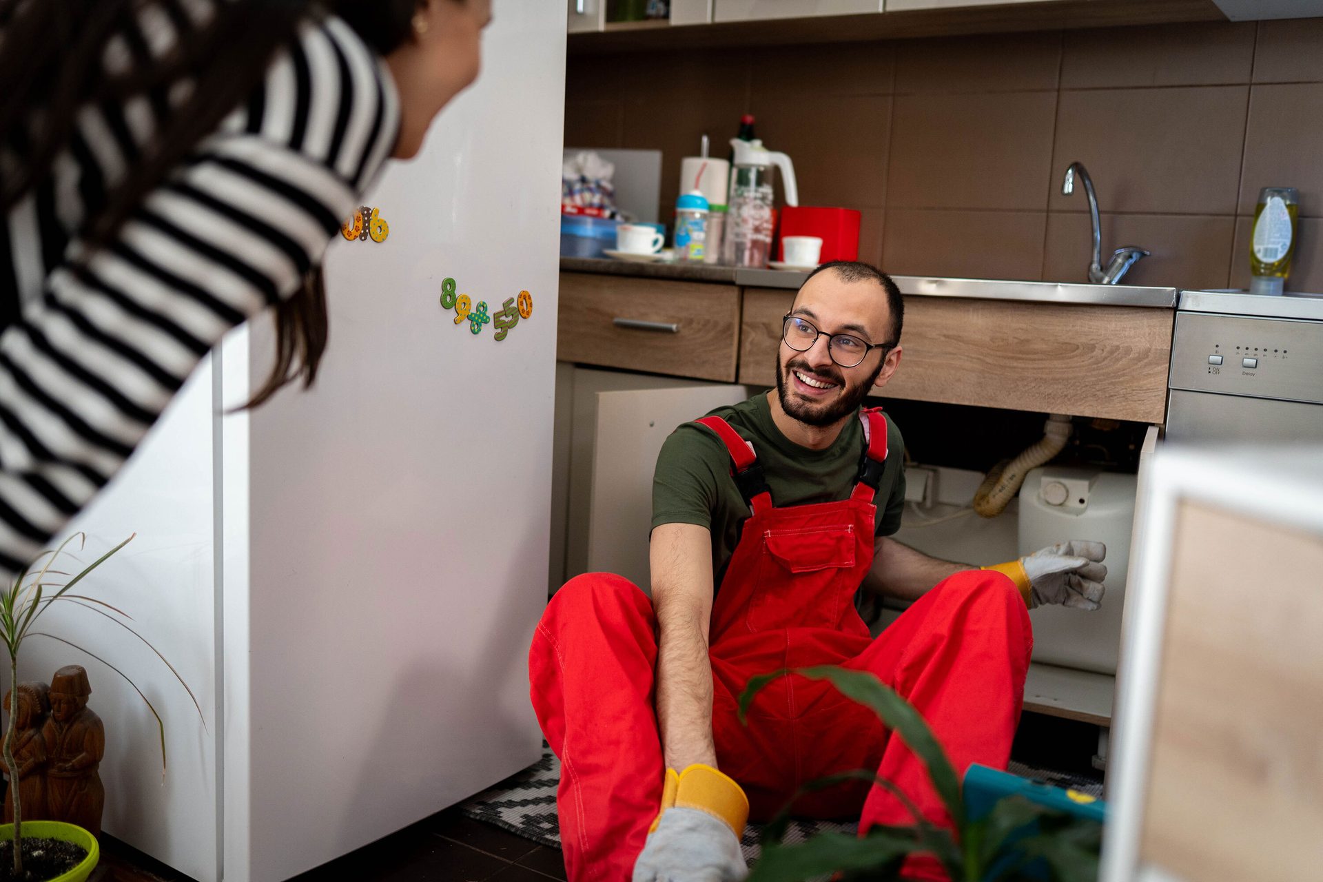 Smiling man in red overalls fixes sink, watched by woman in a kitchen.
