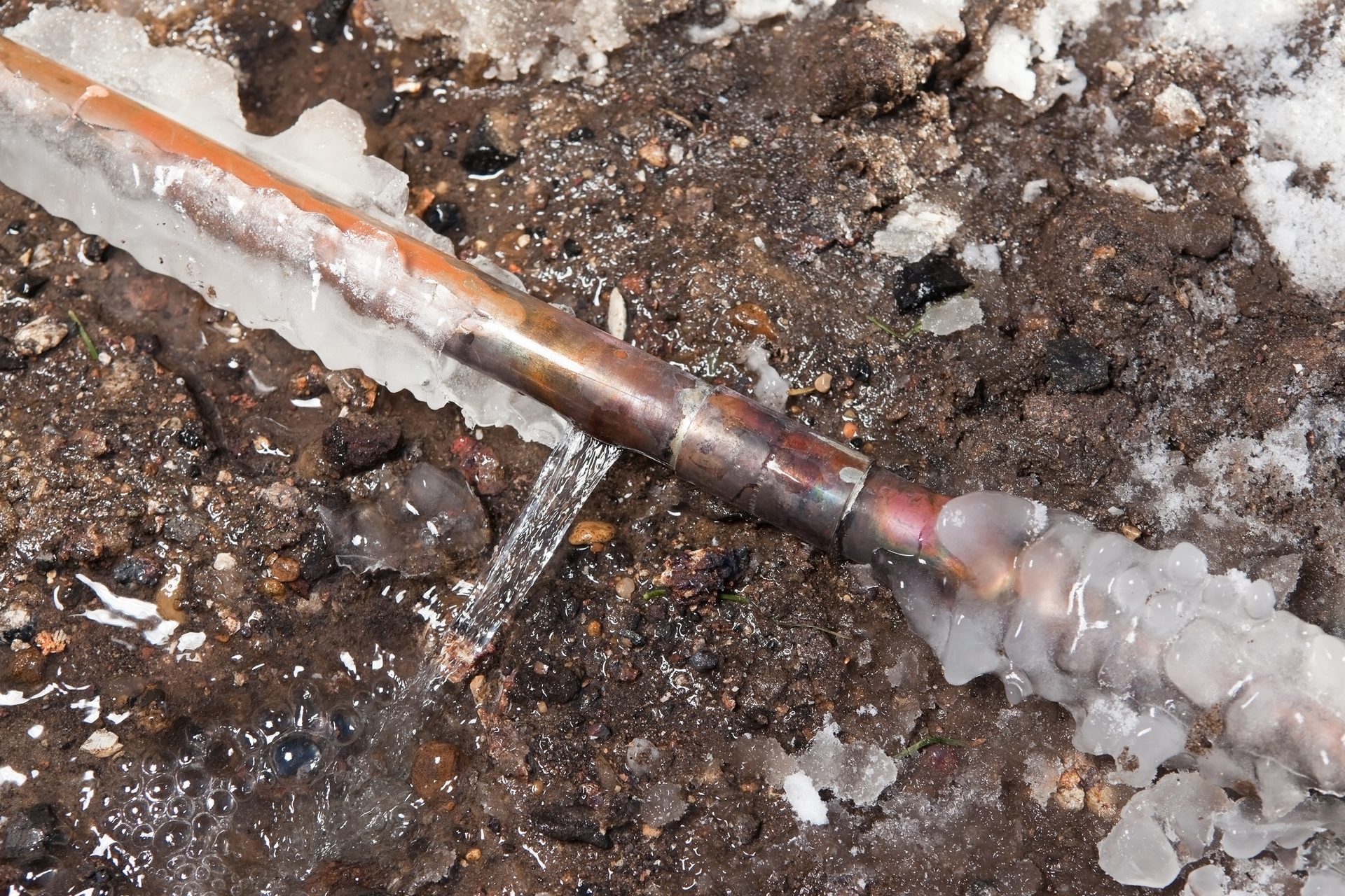 A burst, frozen copper pipe leaking water onto icy, muddy ground.