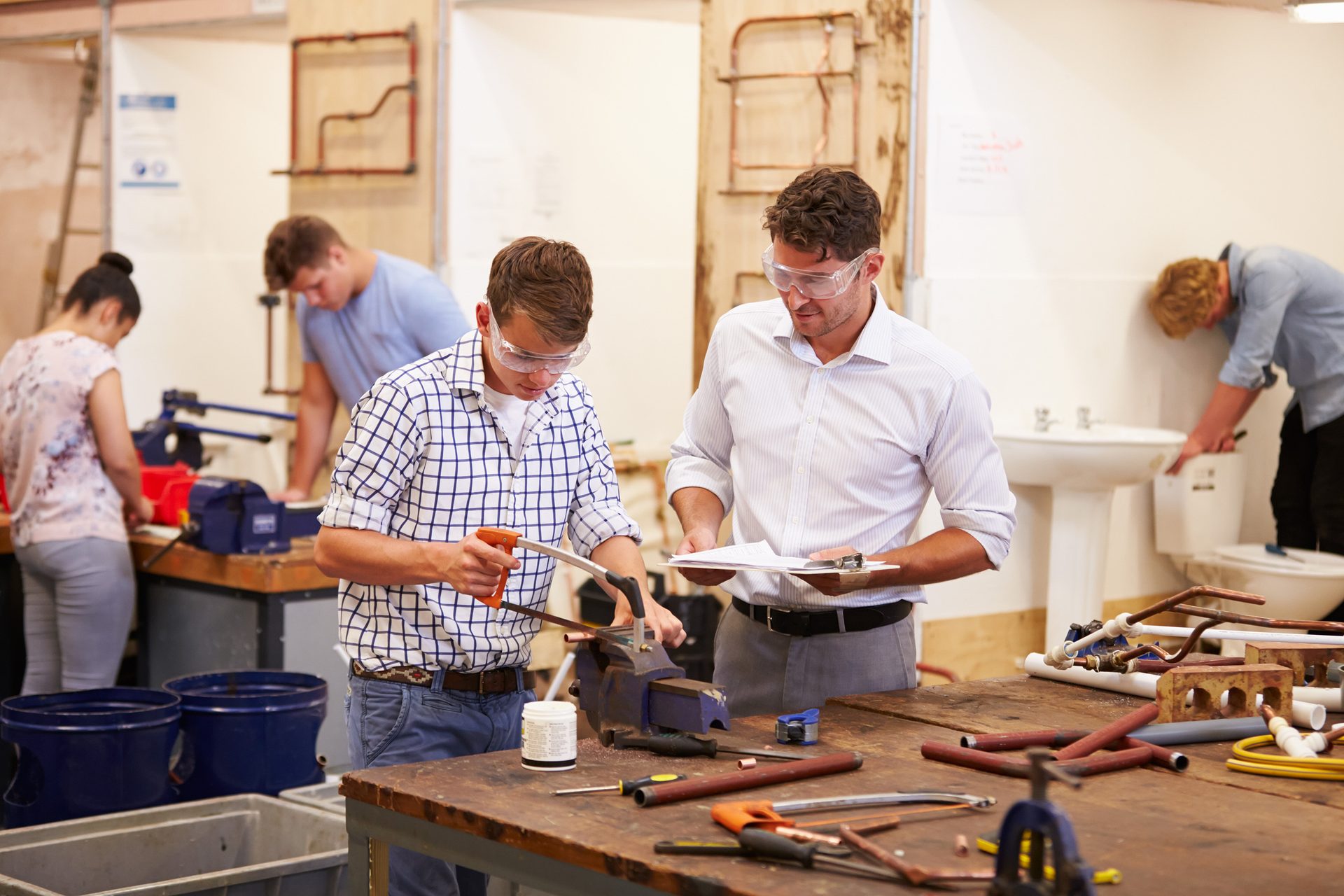 Students and instructor in a trades workshop, wearing safety glasses, working on pipes.