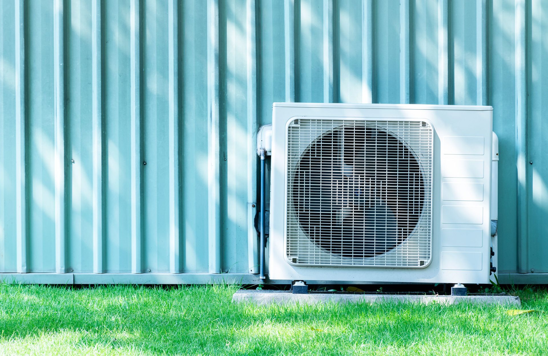 White outdoor AC unit next to a light blue corrugated metal wall and green grass.