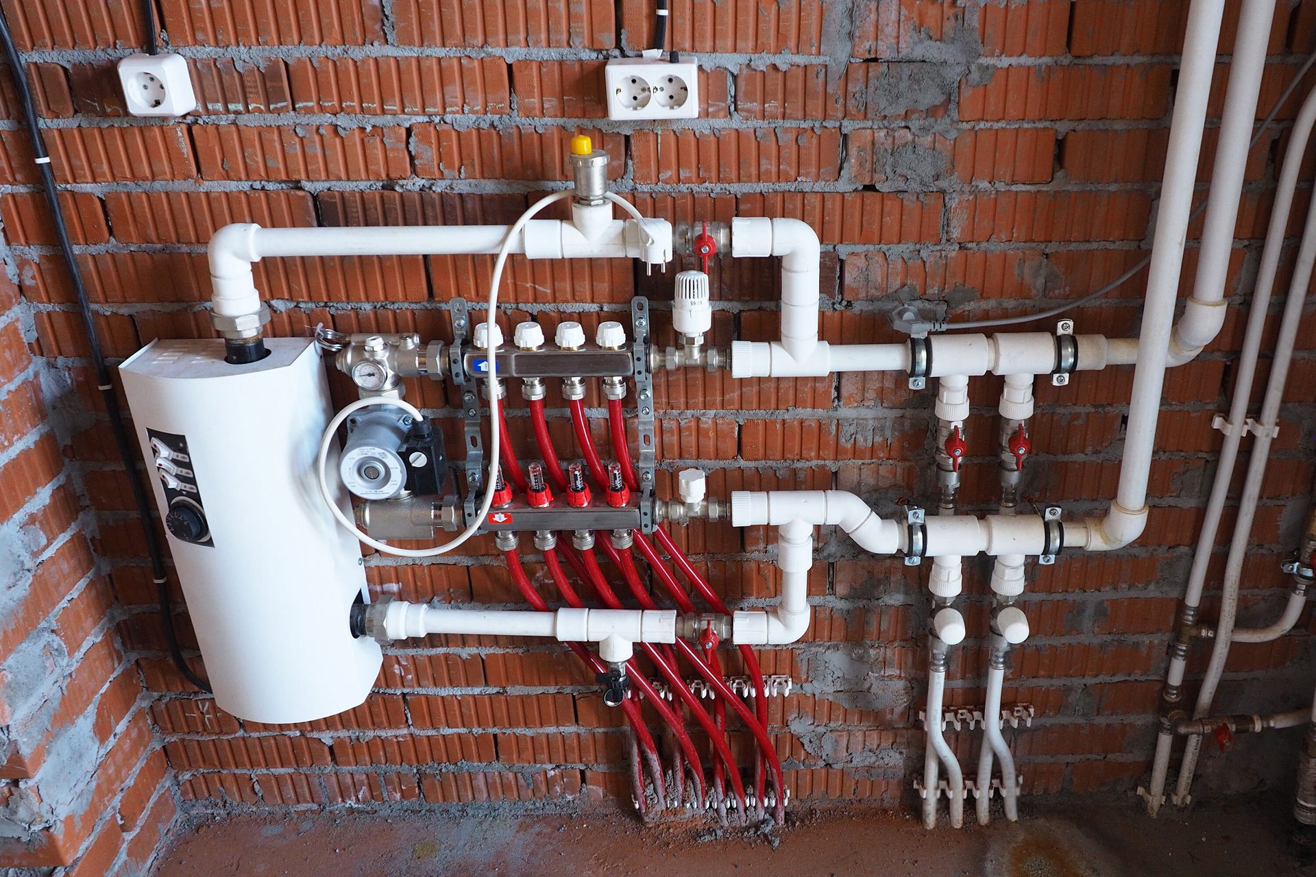 Underfloor heating manifold, boiler, and pipework with red and white pipes on a brick wall.